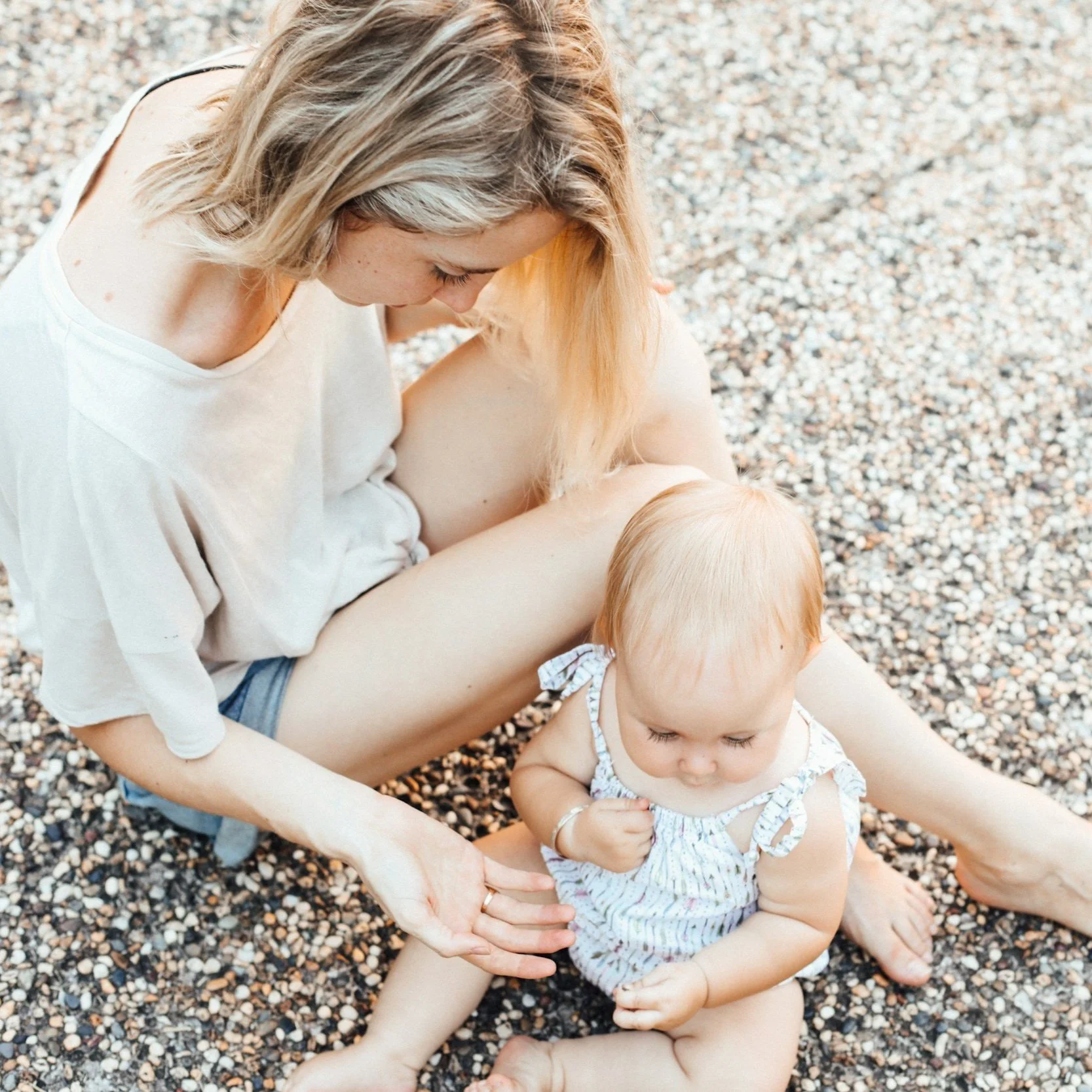 A woman with blonde hair playing in the pebbles with an older infant