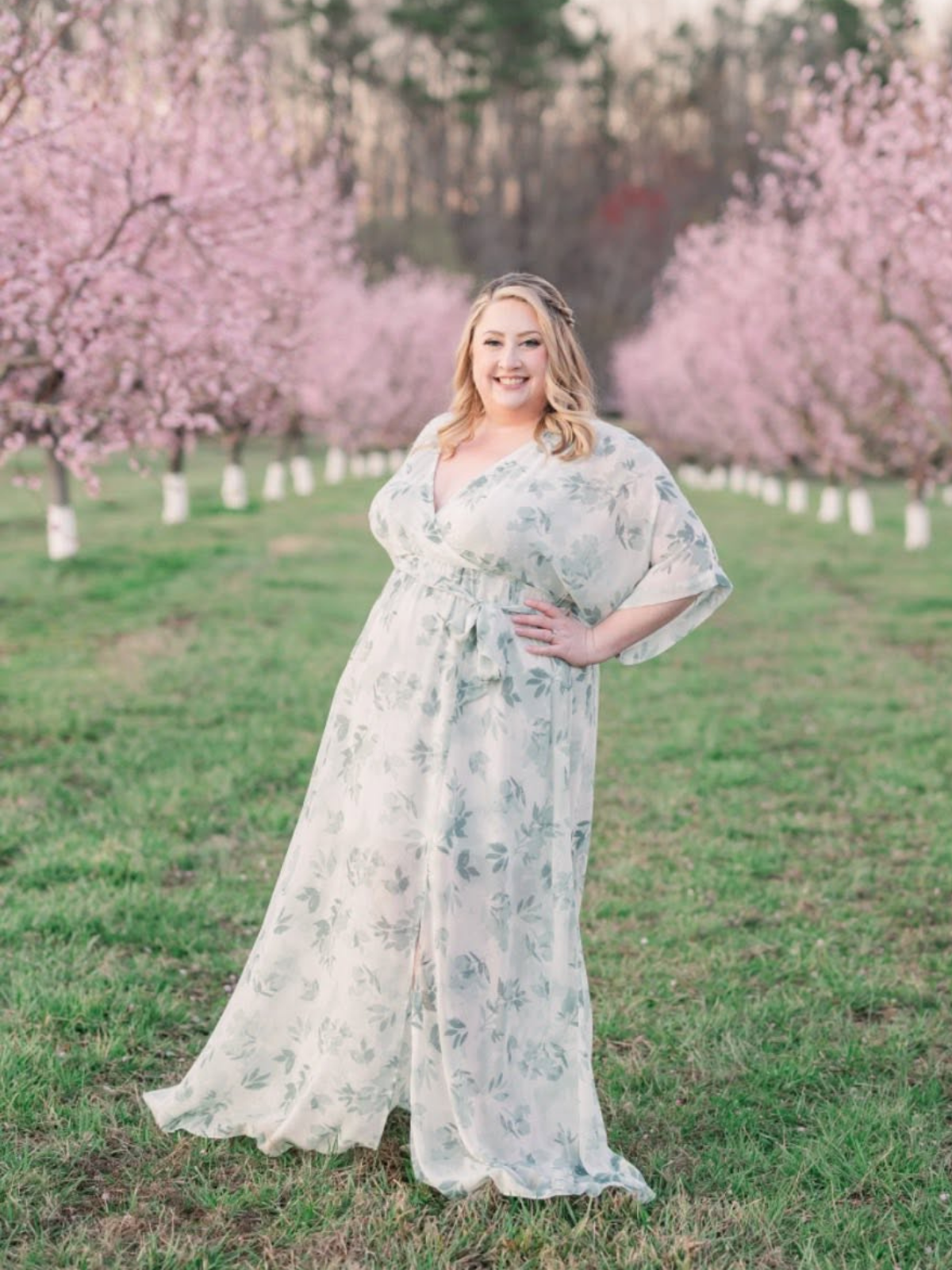 A woman in a floral dress standing in a row of blossoming pink cherry trees during daytime.
