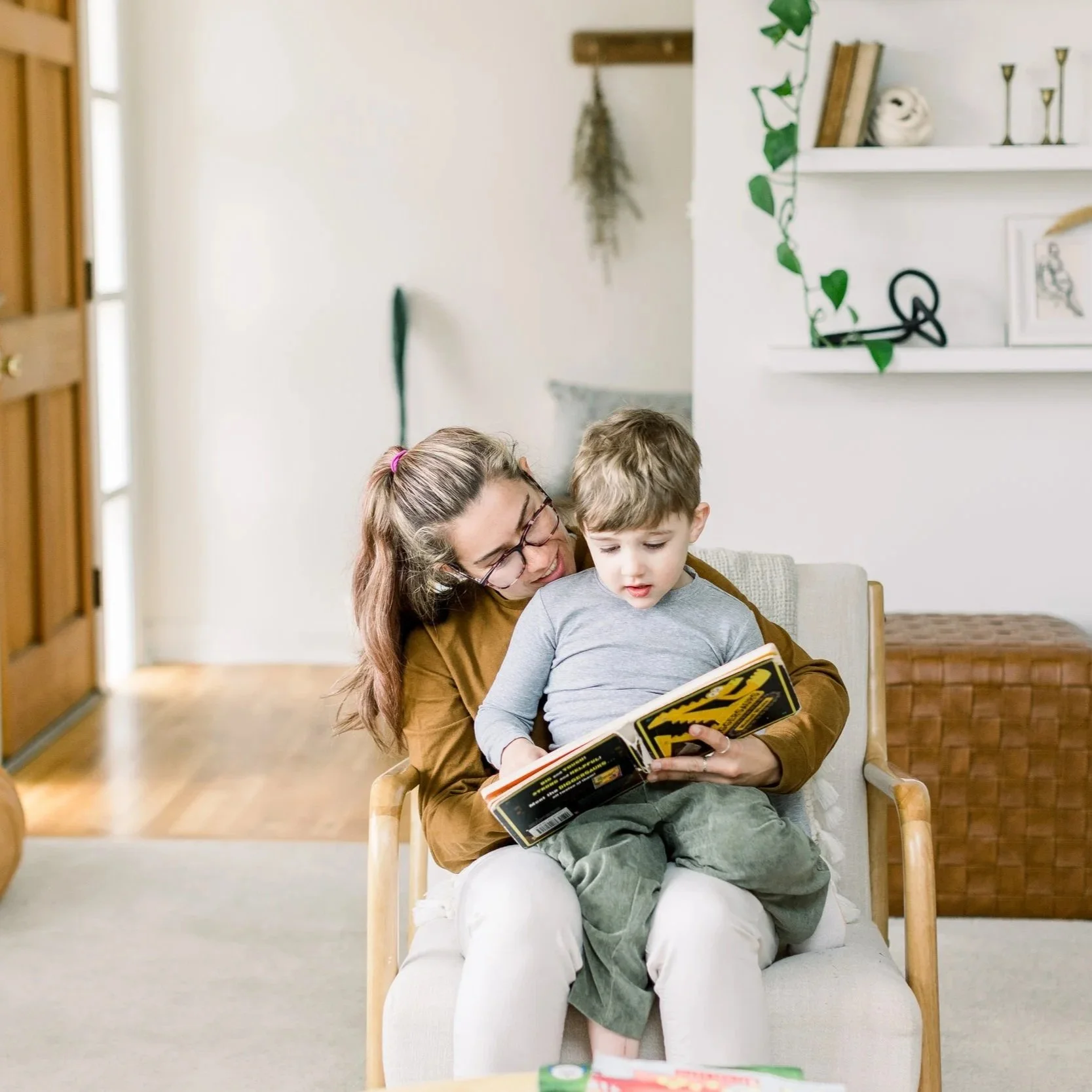 A woman with glasses and long hair tied with a pink hairband sitting on a chair wearing a brown top, holding a young boy on her lap, reading a comic book together in a cozy home interior.