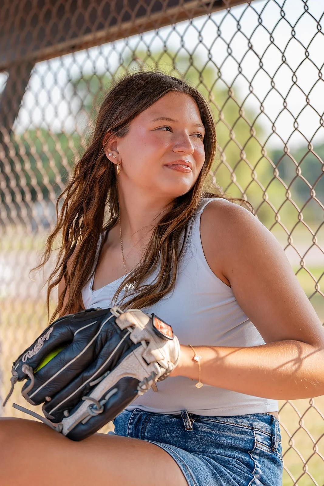 A senior photo  of a young woman with long wavy brown hair, wearing a white tank top and denim shorts, sitting by a chain-link fence holding a baseball glove, with a blurred background of green trees and blue sky.