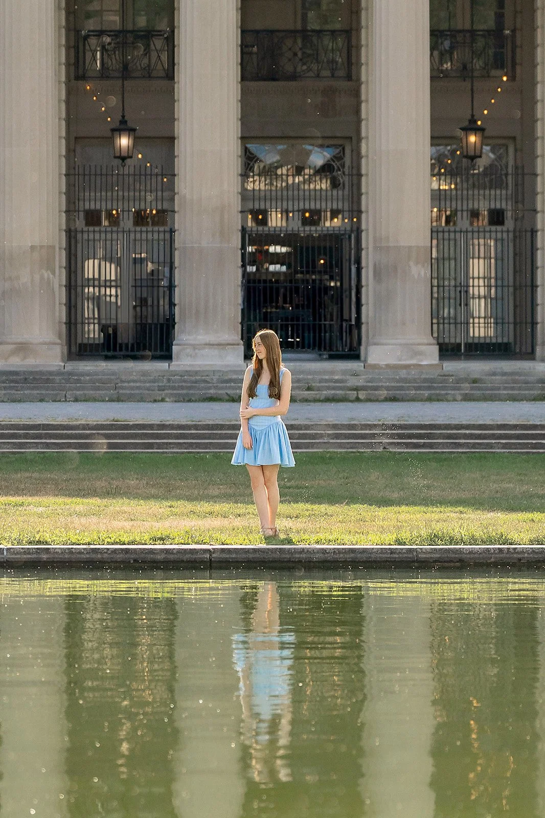 A senior portrait of a young woman in a light blue dress standing on the grass by a body of water, with a large building with columns and iron gates in the background.