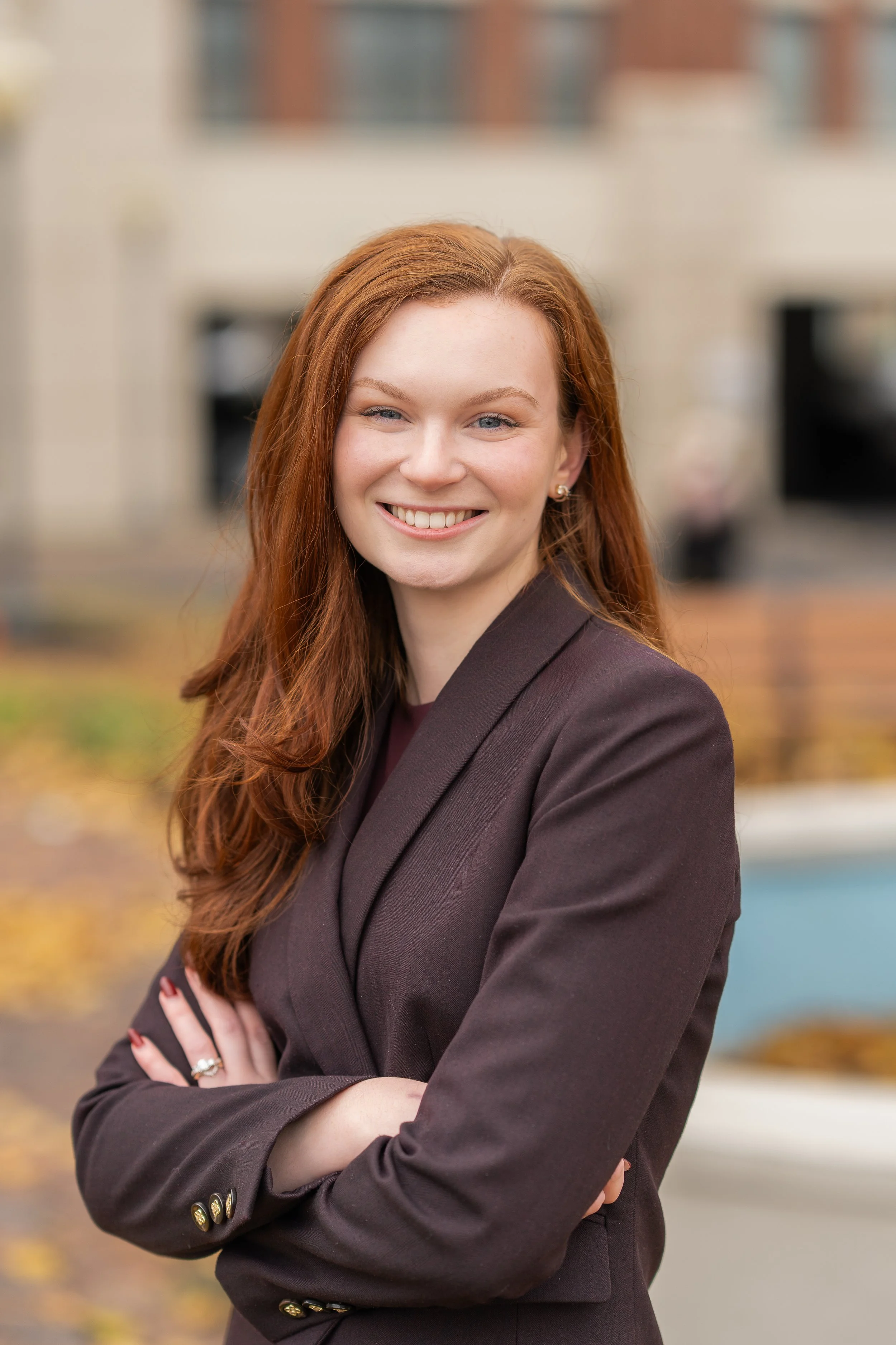 Headshot photo of a woman with red hair and blue eyes smiling outdoors in front of a blurred background with autumn leaves and a building.