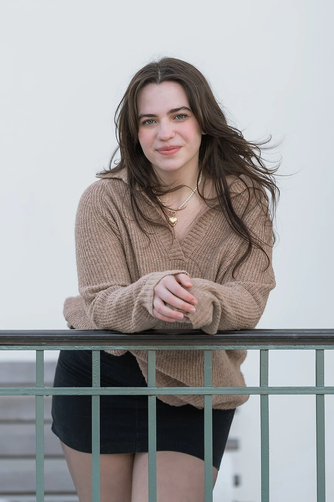 A senior photo of a young woman with dark hair and blue eyes wearing a beige sweater and black shorts, leaning on a metal railing, smiling at the camera.