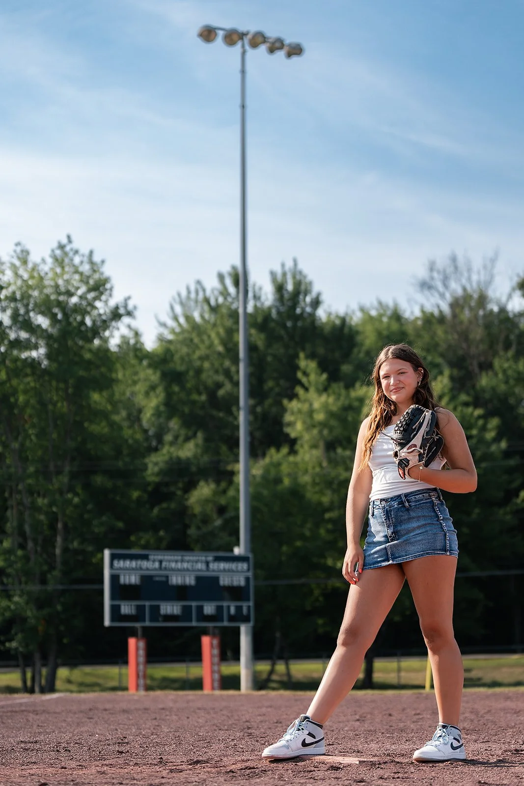 Senior photo of a young woman standing on a baseball field, holding a baseball glove, with a scoreboard and trees in the background.