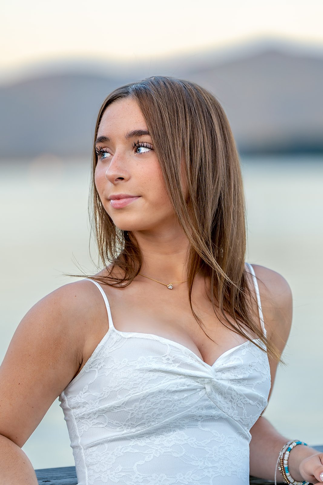 Senior portrait of a young woman with brown hair and blue eyes wearing a white tank top, standing outdoors near water with mountains in the background.