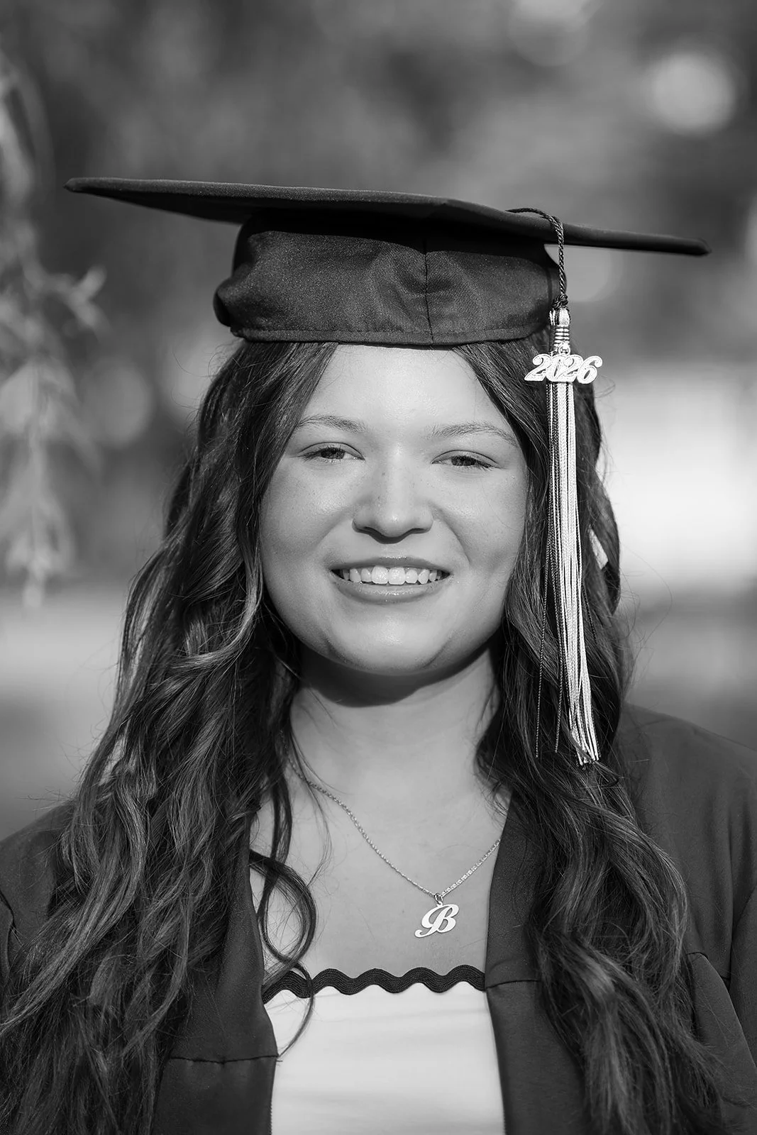 Black and white photo of a young woman celebrating her graduation, wearing a cap with a "2026" tassel, smiling, with long wavy hair, and wearing a necklace with the letter 'B'.