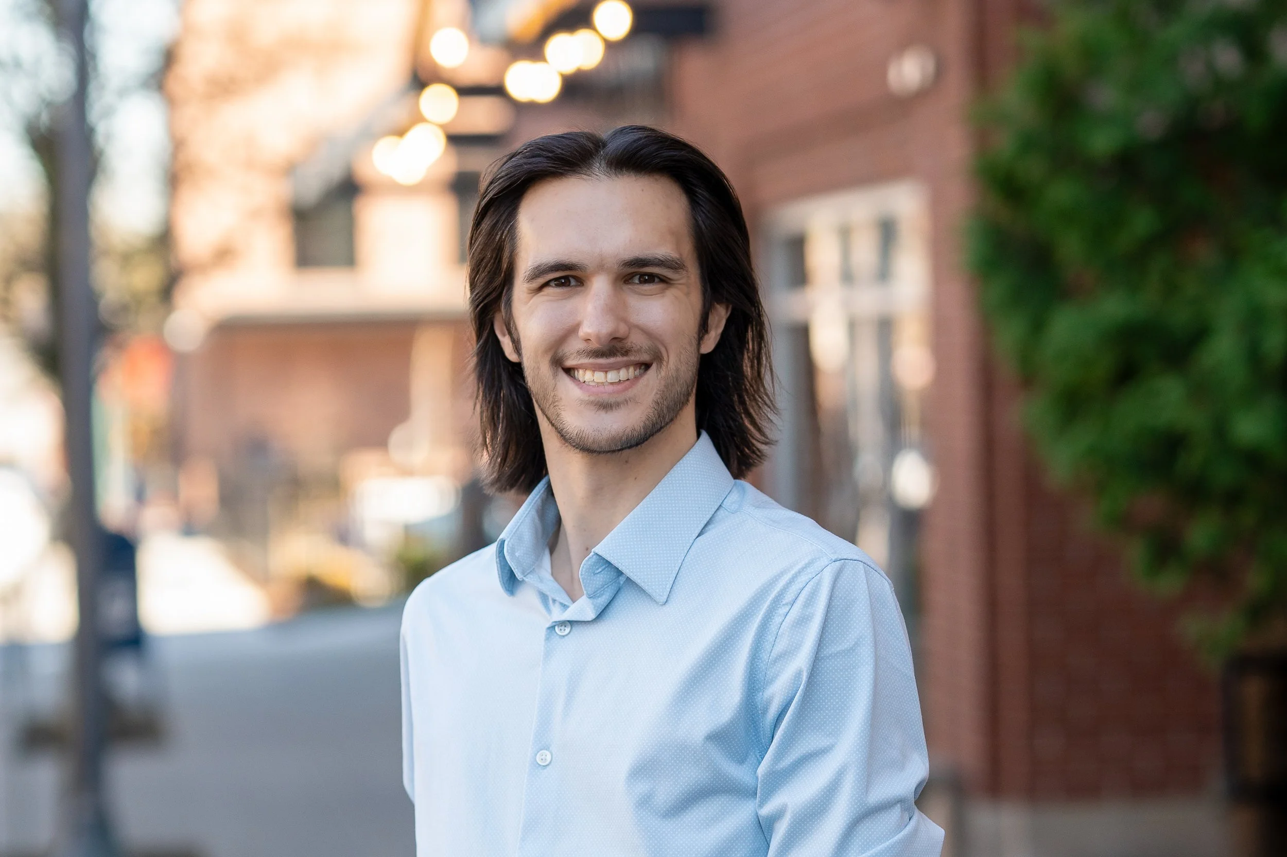 A headshot of a young man with dark, shoulder-length hair, wearing a light blue dress shirt, standing outdoors in front of a blurred background of buildings, trees, and warm lighting.