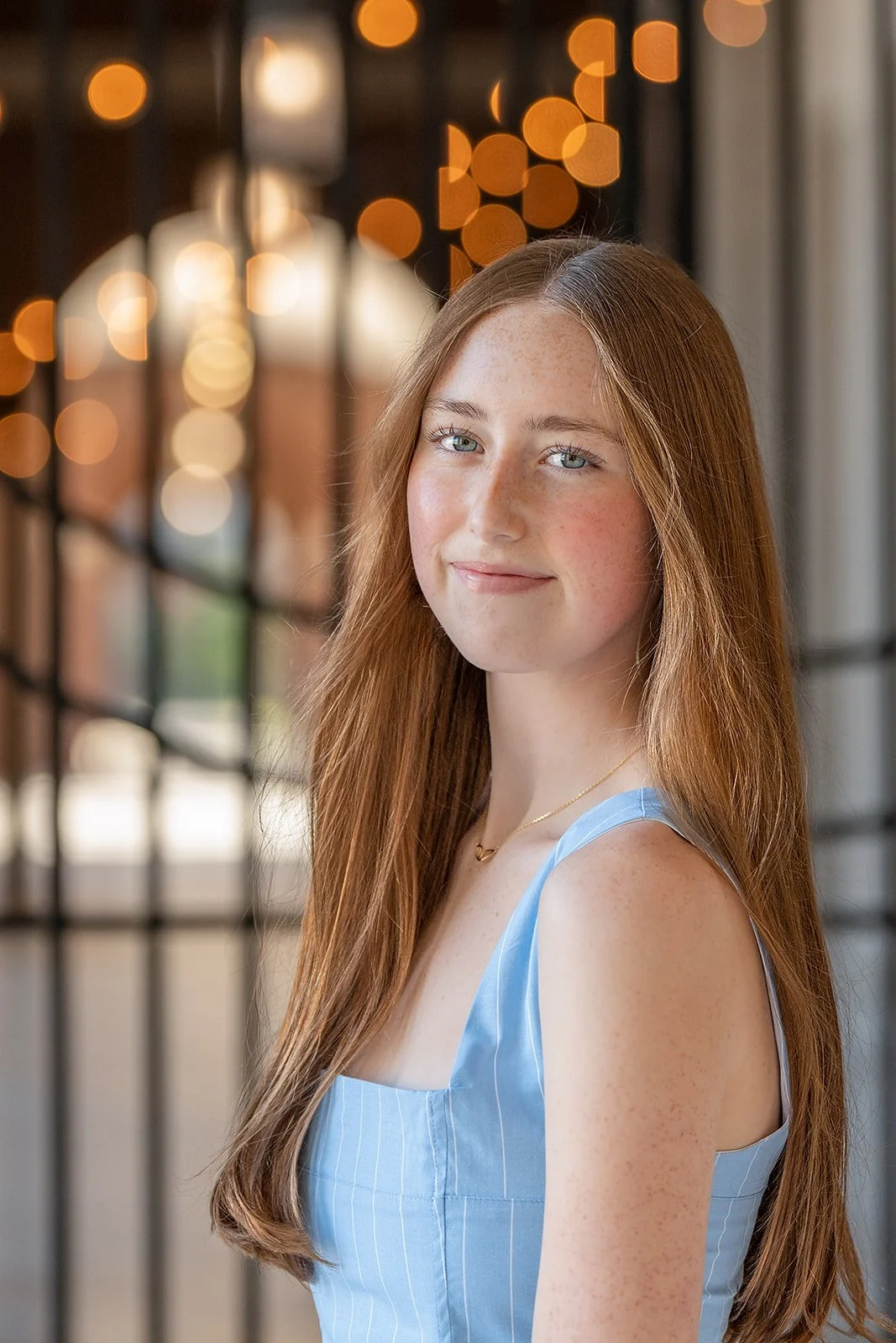 A senior portrait of a young woman with long red hair and clear skin wearing a light blue sleeveless top, smiling lightly, standing indoors with blurred warm-toned hanging lights in the background.