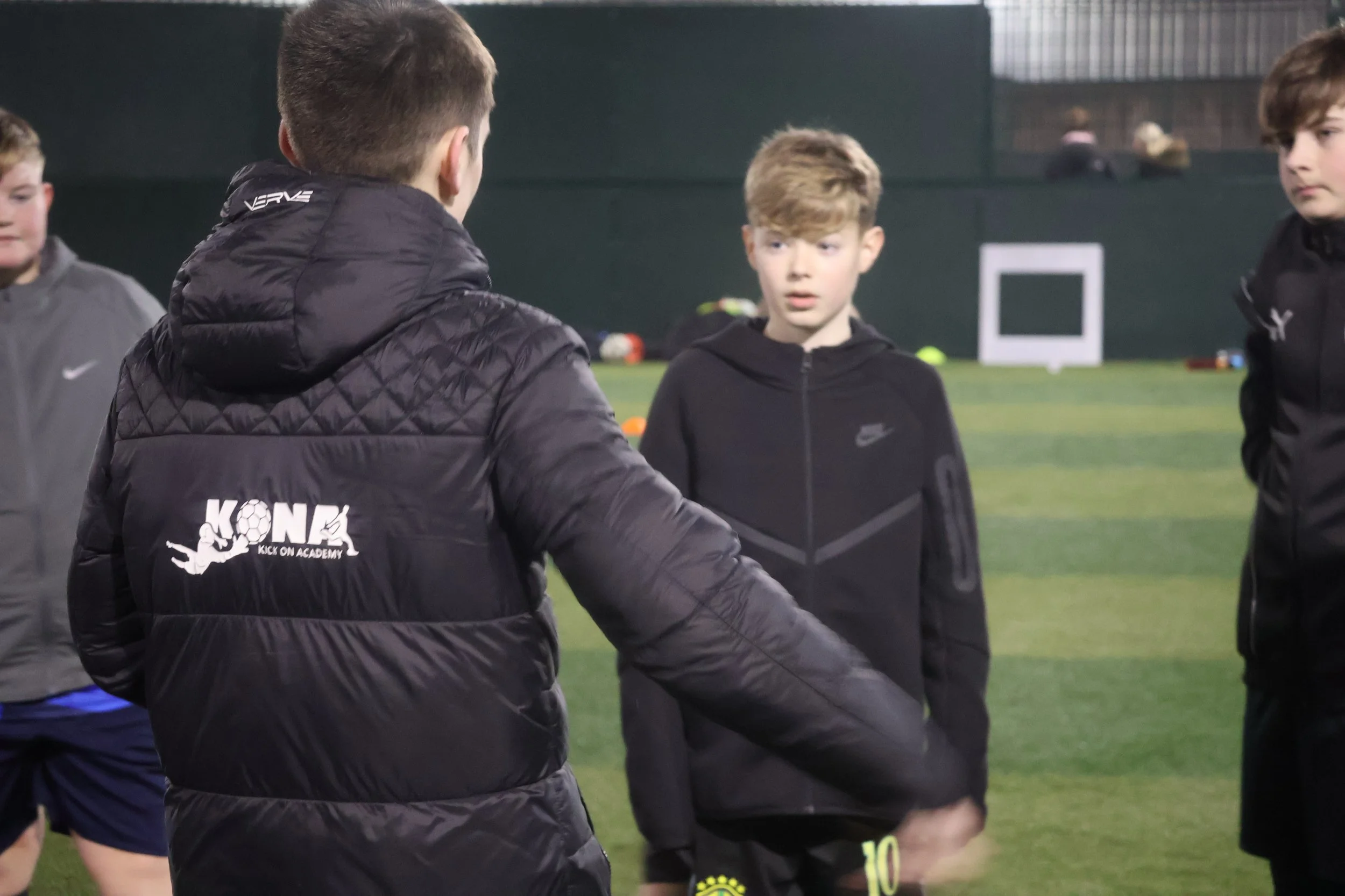 A coach in a black jacket talking to young soccer players on a field at night, with soccer balls and a goal in the background.