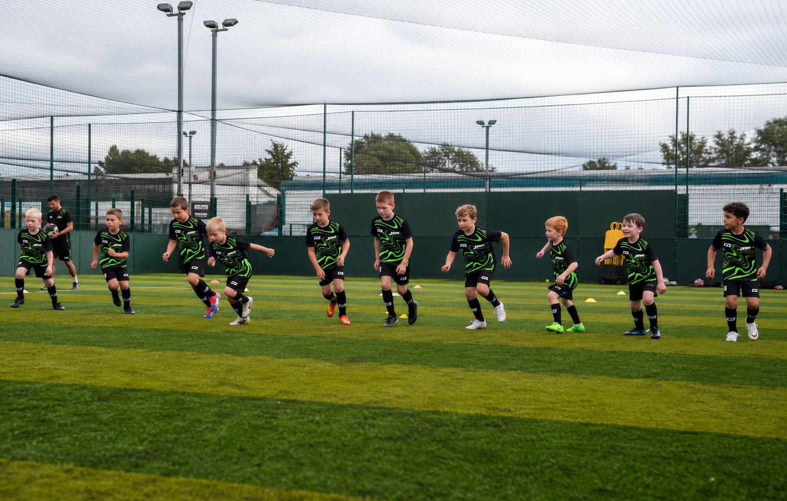 Children in black and green soccer uniforms are practicing on a field, with a netted fence and stadium lights in the background.