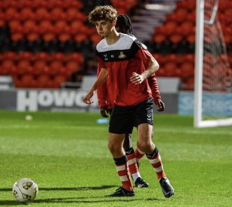 A young soccer player on the field during a practice or warm-up, with a soccer ball nearby, wearing a black and red jersey and striped red and white socks.