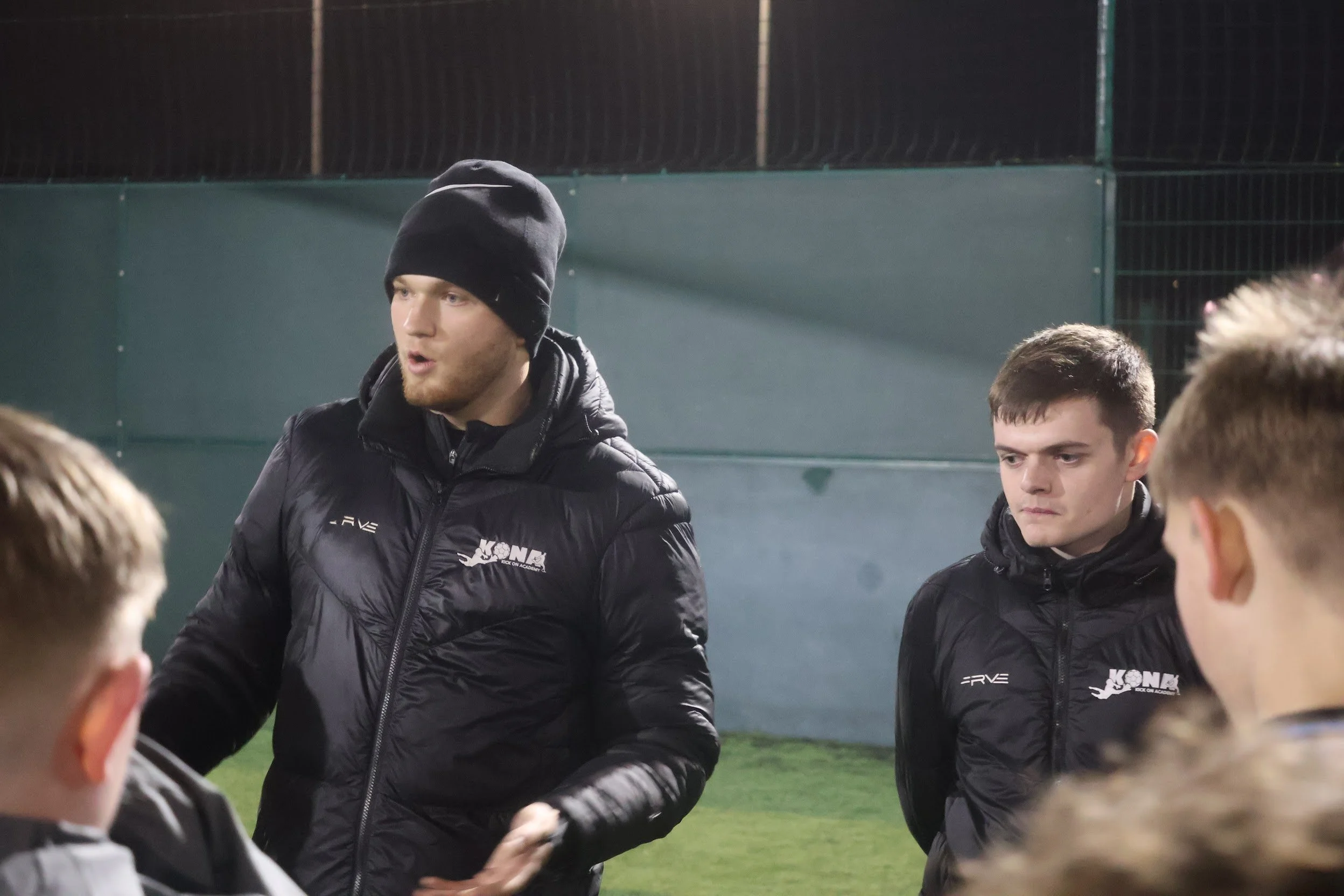 Two young men wearing black jackets and beanies are standing outdoors, speaking to a group of people during nighttime; the background shows a green wall and a chain-link fence.