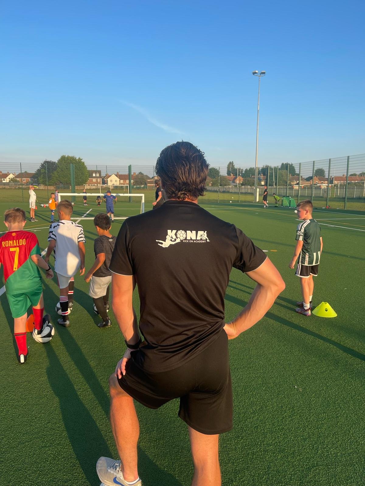 Soccer coach overseeing a youth soccer practice on an outdoor field, children lining up for drills, with a clear blue sky and houses in the background.