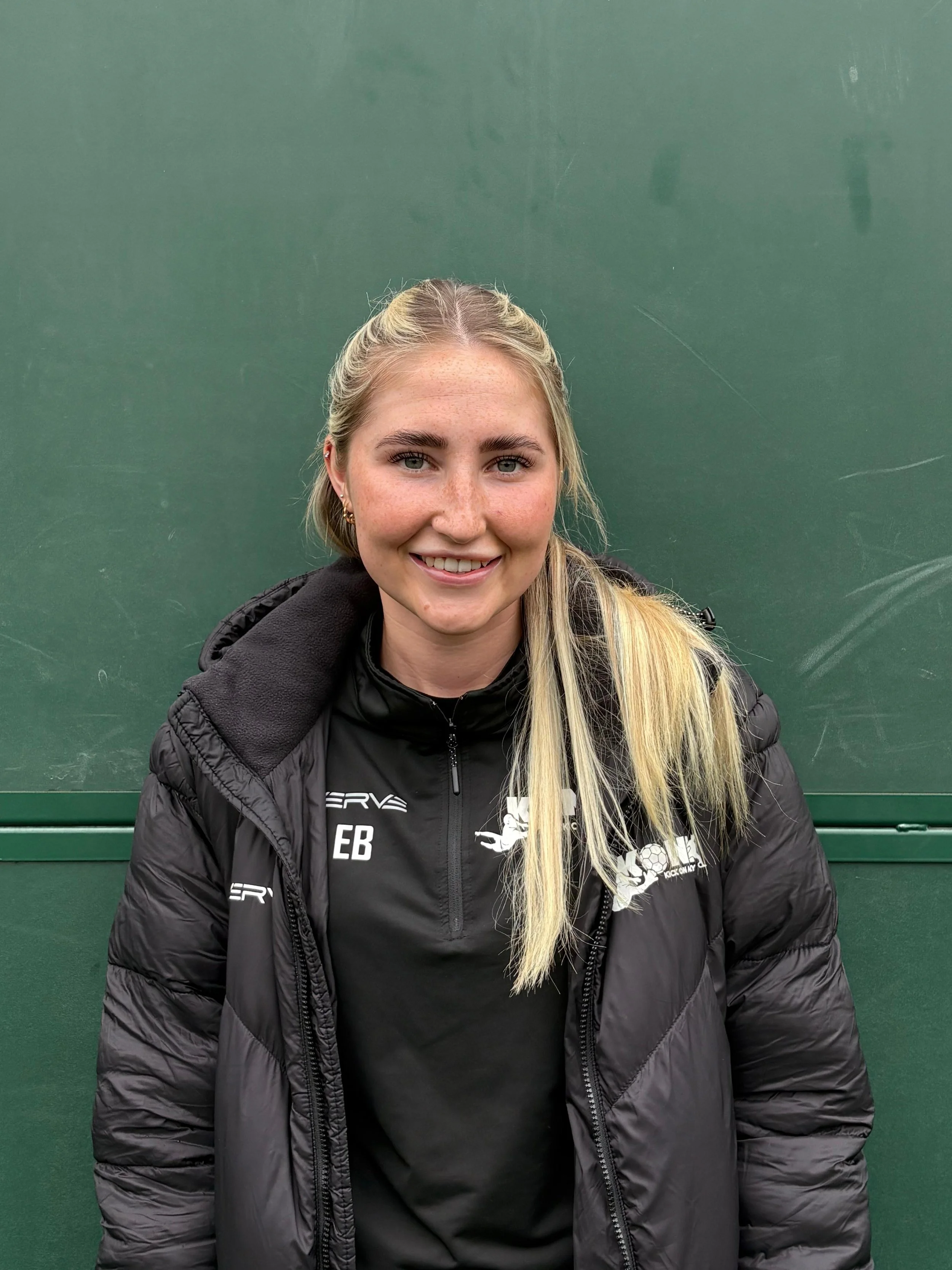 A young woman with long blonde hair, wearing a black sports jacket with a logo and text on it, smiling in front of a green chalkboard or wall.