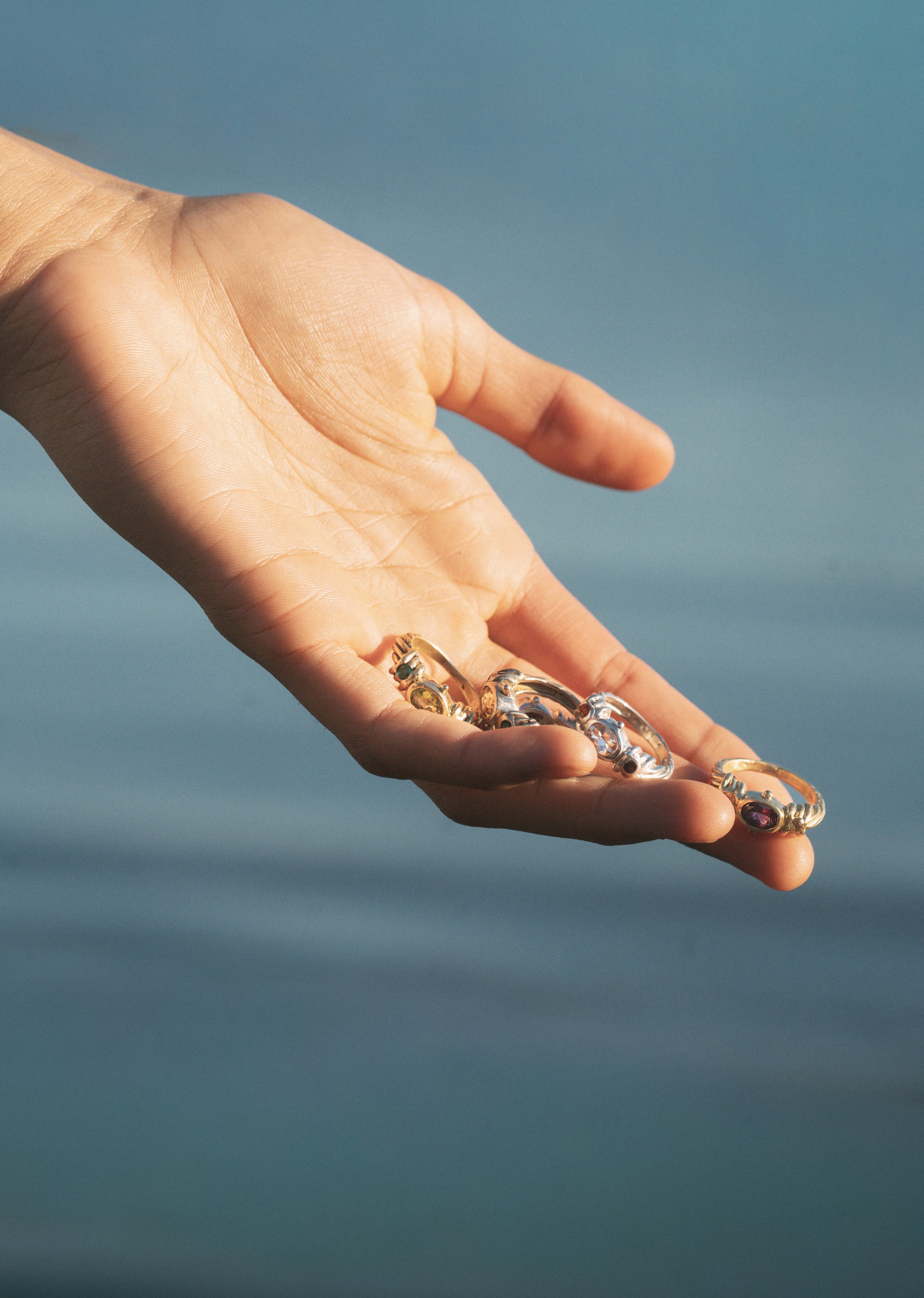 A hand holding several rings with different gemstones against a blue water background.