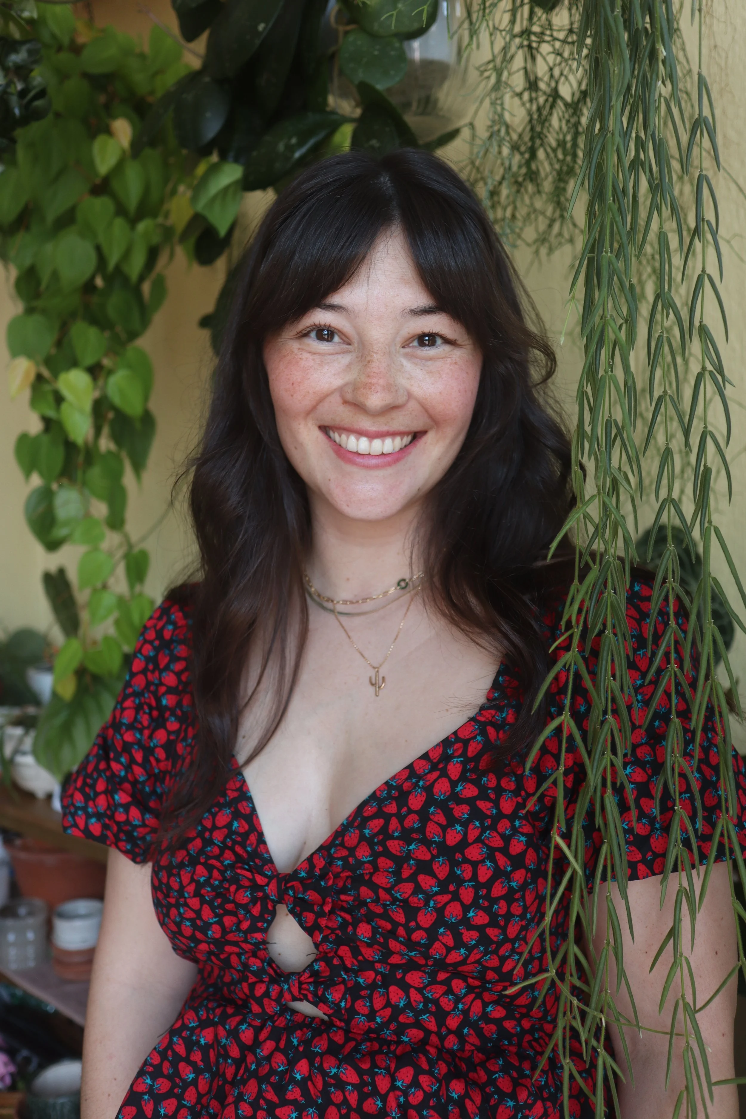 A smiling woman with long, wavy dark hair, wearing a black dress with a red strawberry pattern, jewelry including necklaces, standing among green leafy plants.