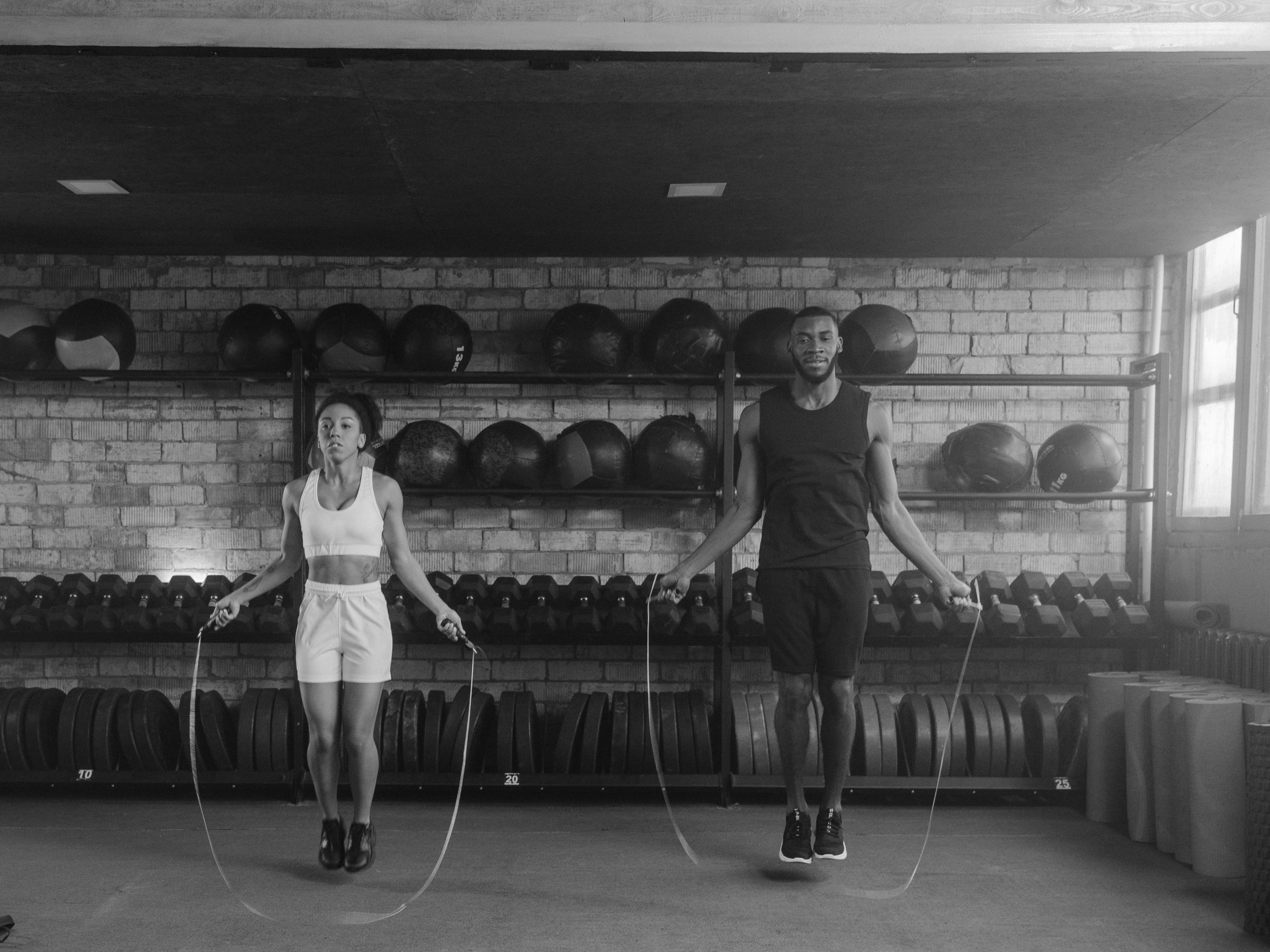 Two people jump rope in a gym with brick walls and shelves of medicine balls, dumbbells, and workout equipment.