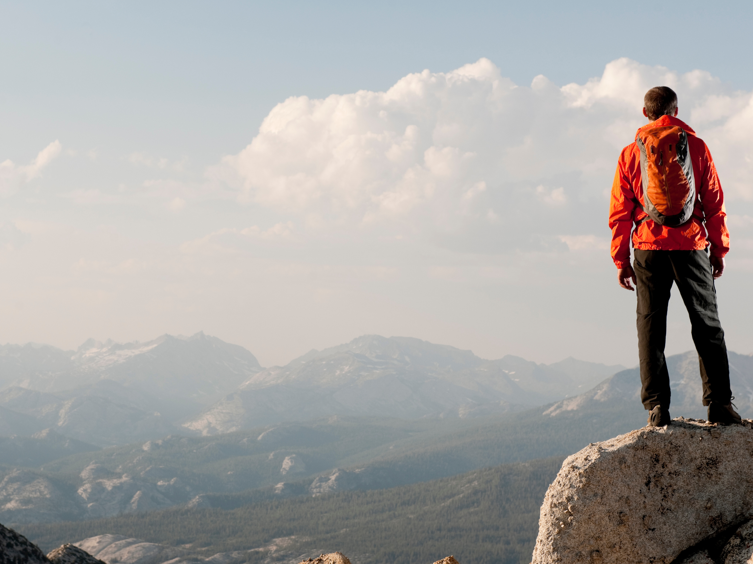 Man standing on a large rock overlooking mountain range, wearing an orange jacket and backpack.