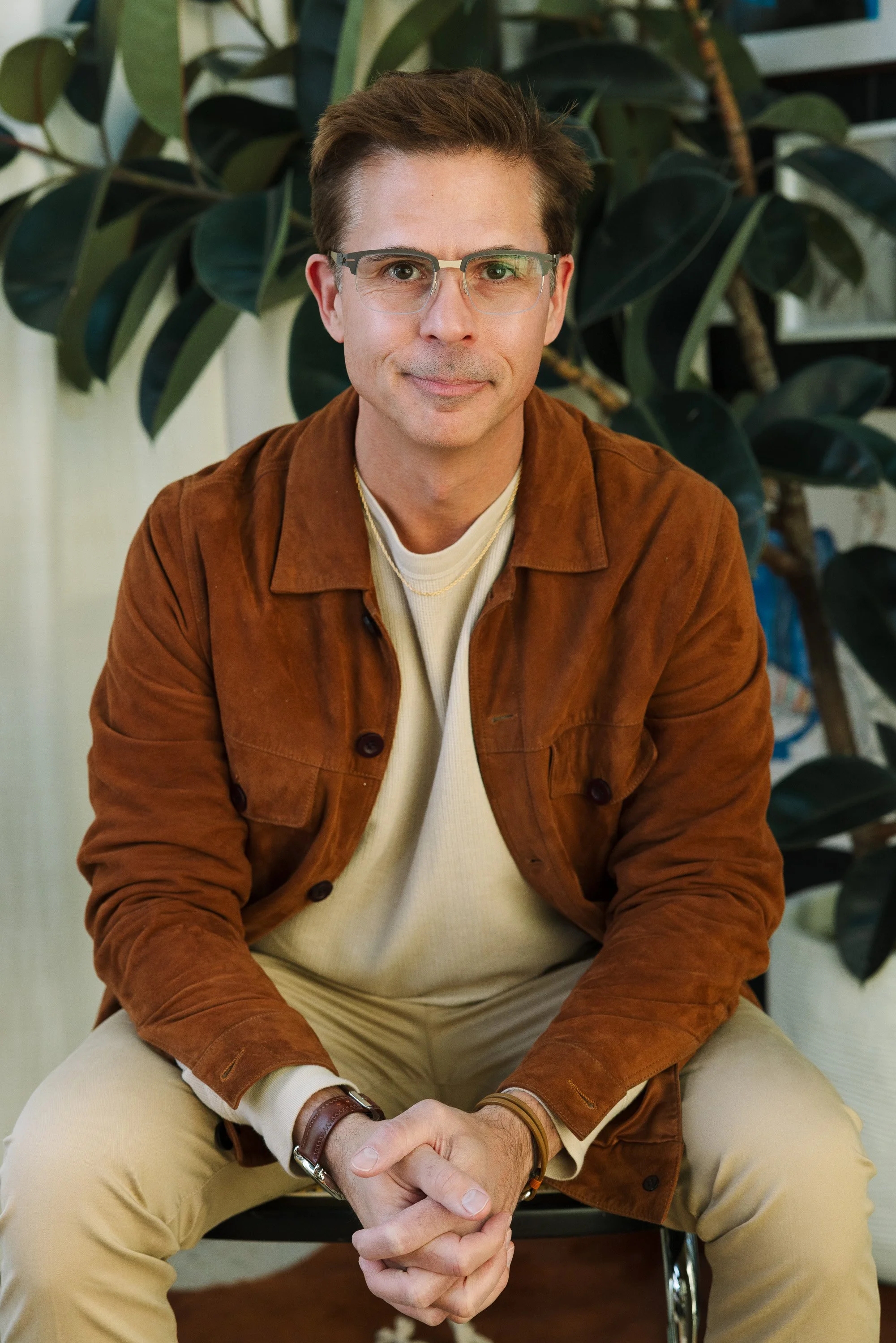 A man with glasses sitting on a chair with hands clasped, wearing a brown jacket over a white shirt, with large green leafy plants in the background.