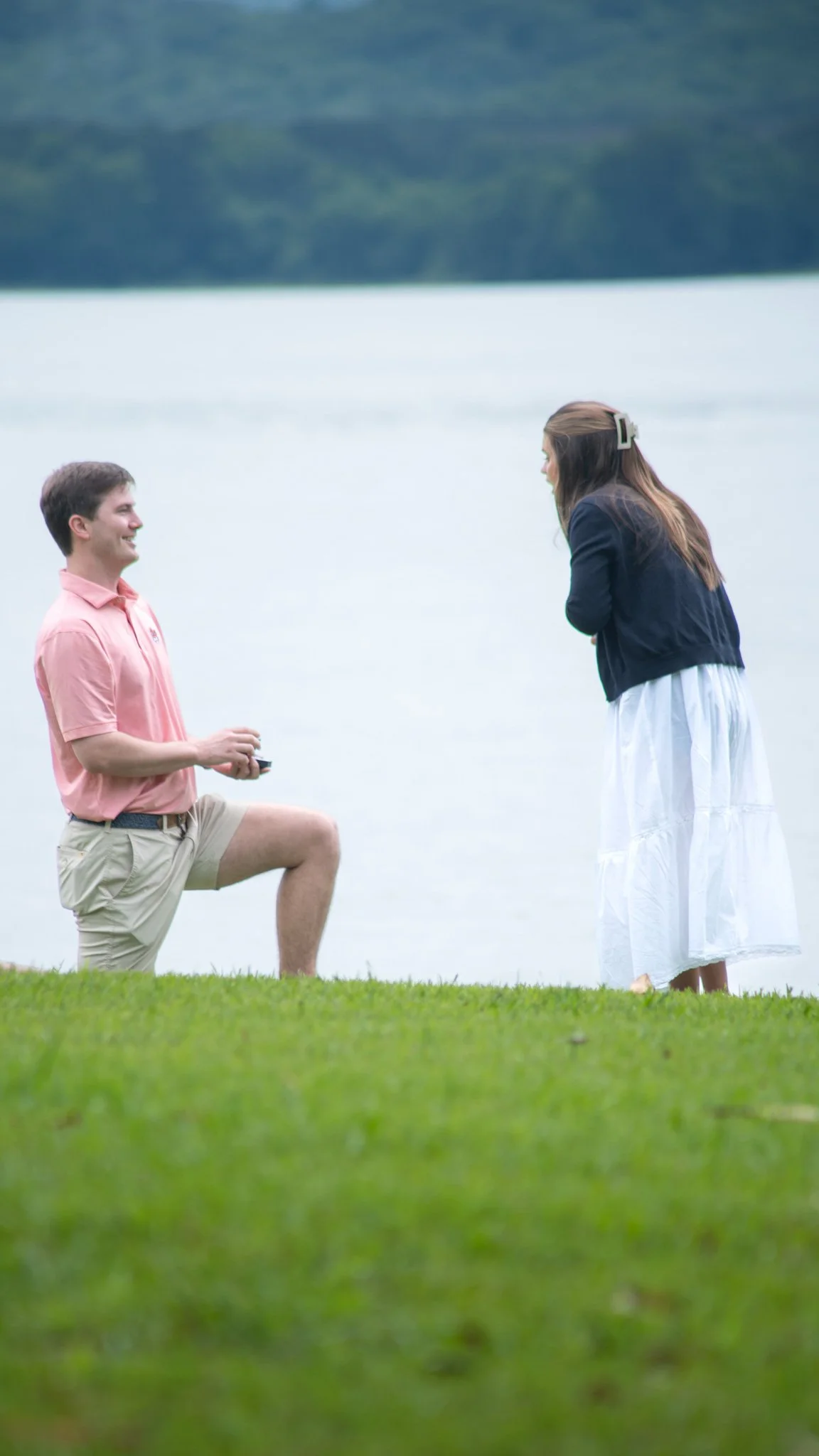 A man kneeling on one knee with a ring box, proposing to a woman near a lake or river with mountains in the background.