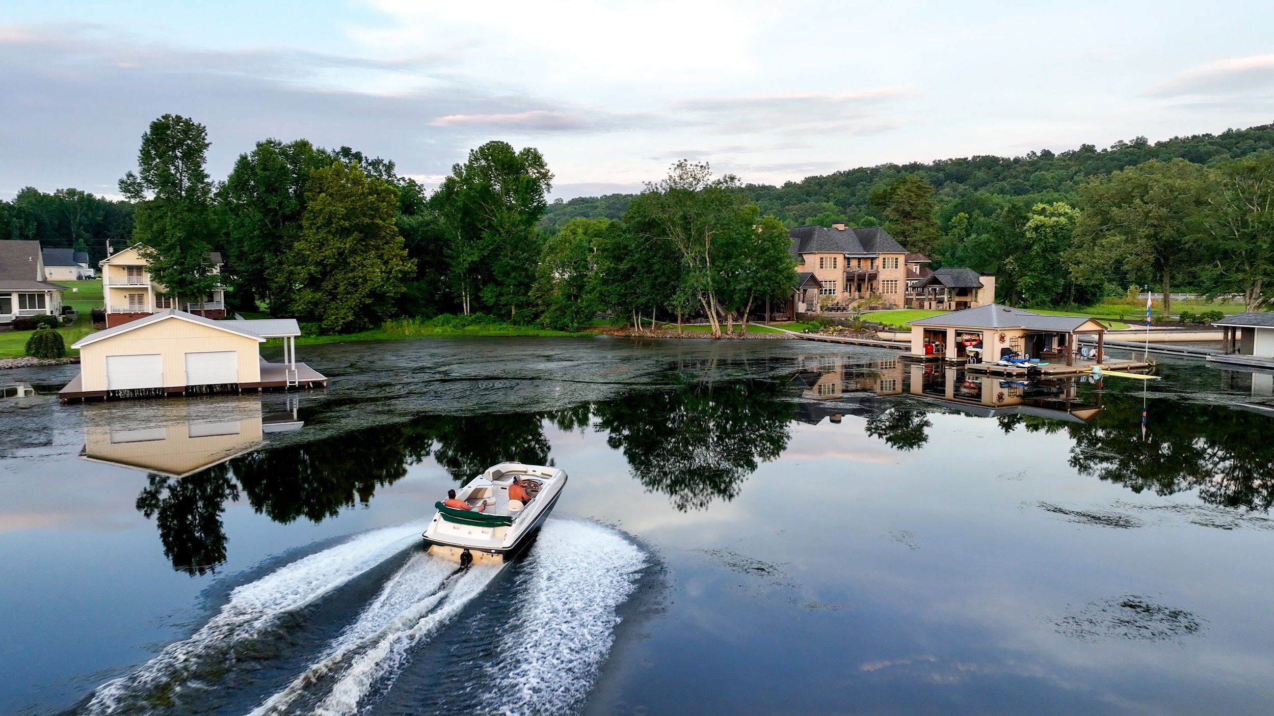 A boat speeds through calm water near a residential lakeside community, with houses, trees, and a boathouse reflected in the water under a partly cloudy sky.