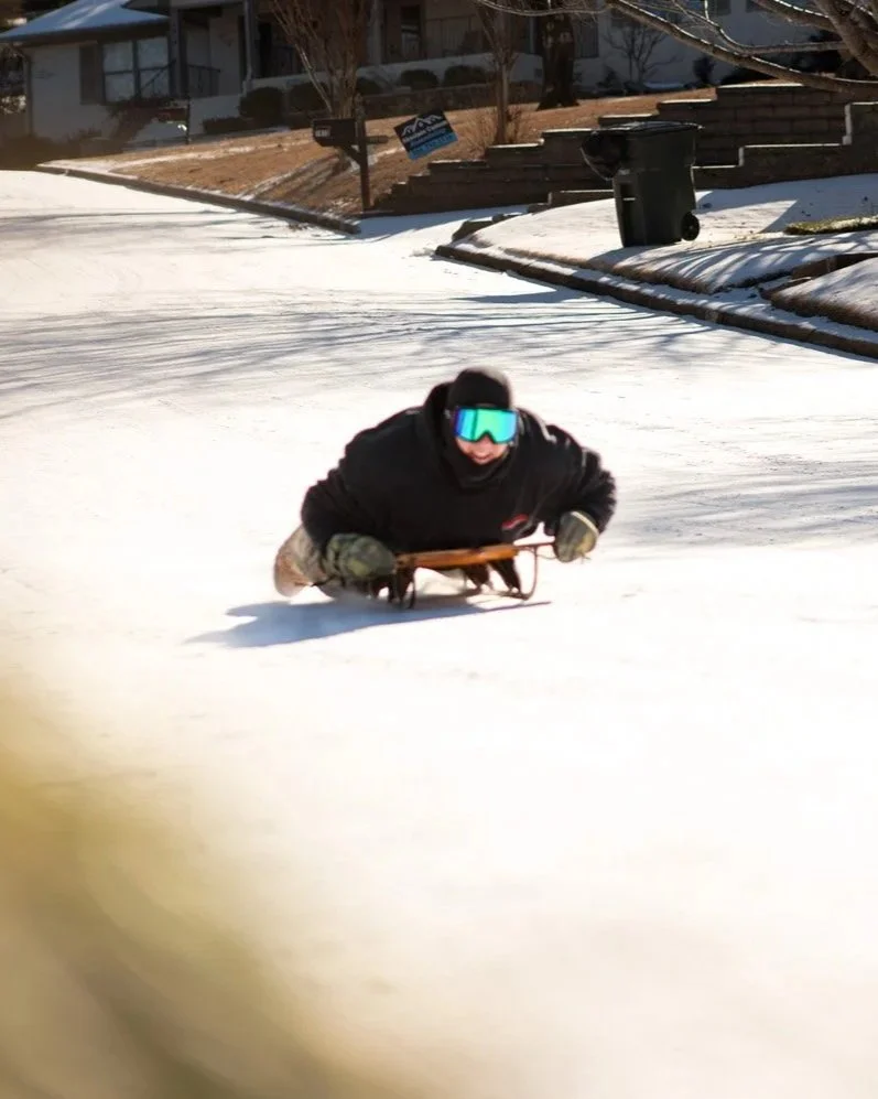 Person in winter clothing and goggles sledding on a snow-covered street.