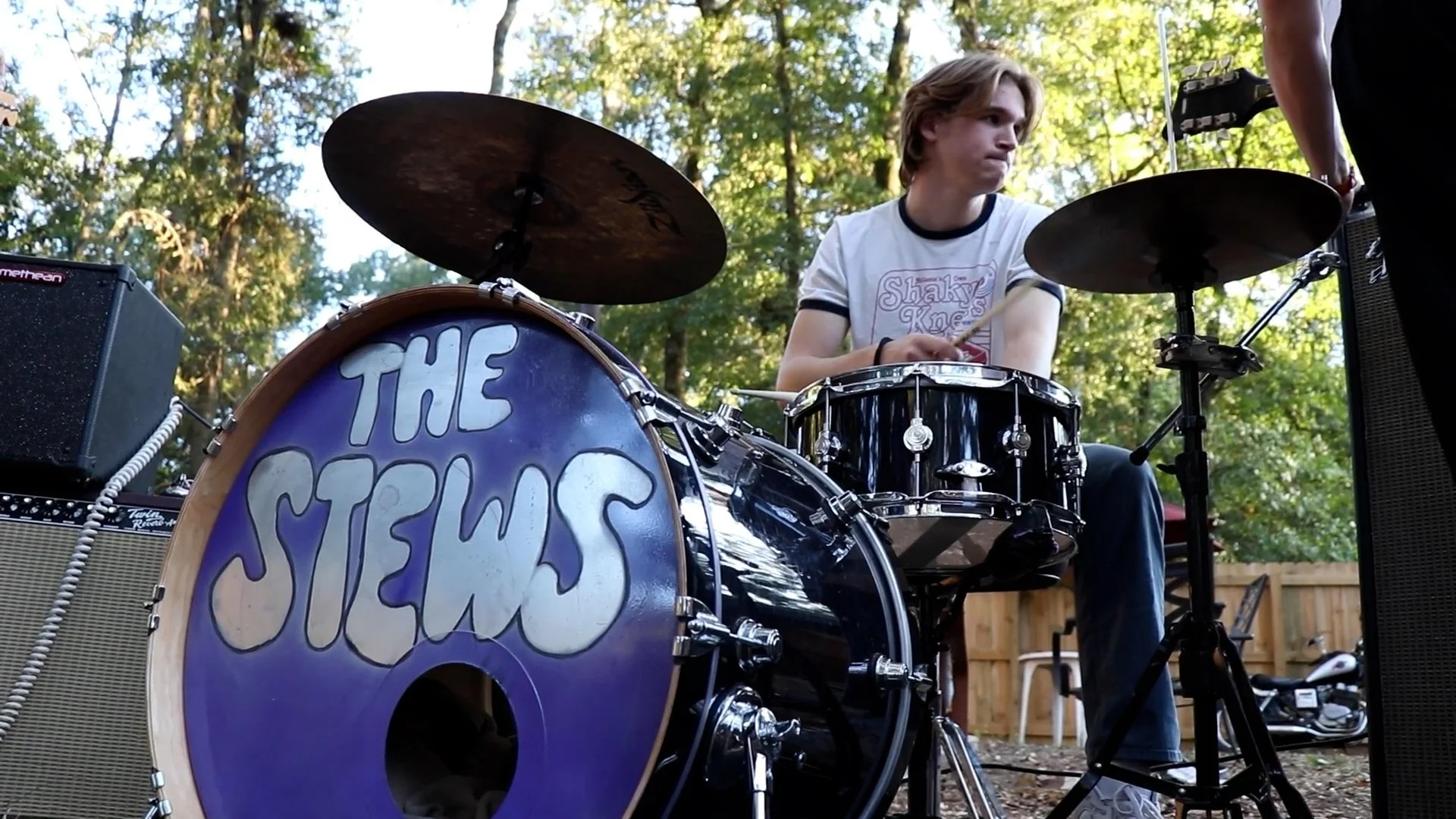 A young man with blond hair playing drums outdoors during the daytime. There is a drum set with the front bass drum displaying the band name 'The Stems' in white text on a purple background. Surrounding the drummer are amplifiers and other musical equipment. The background shows trees and a wooden fence, indicating a backyard or park setting.
