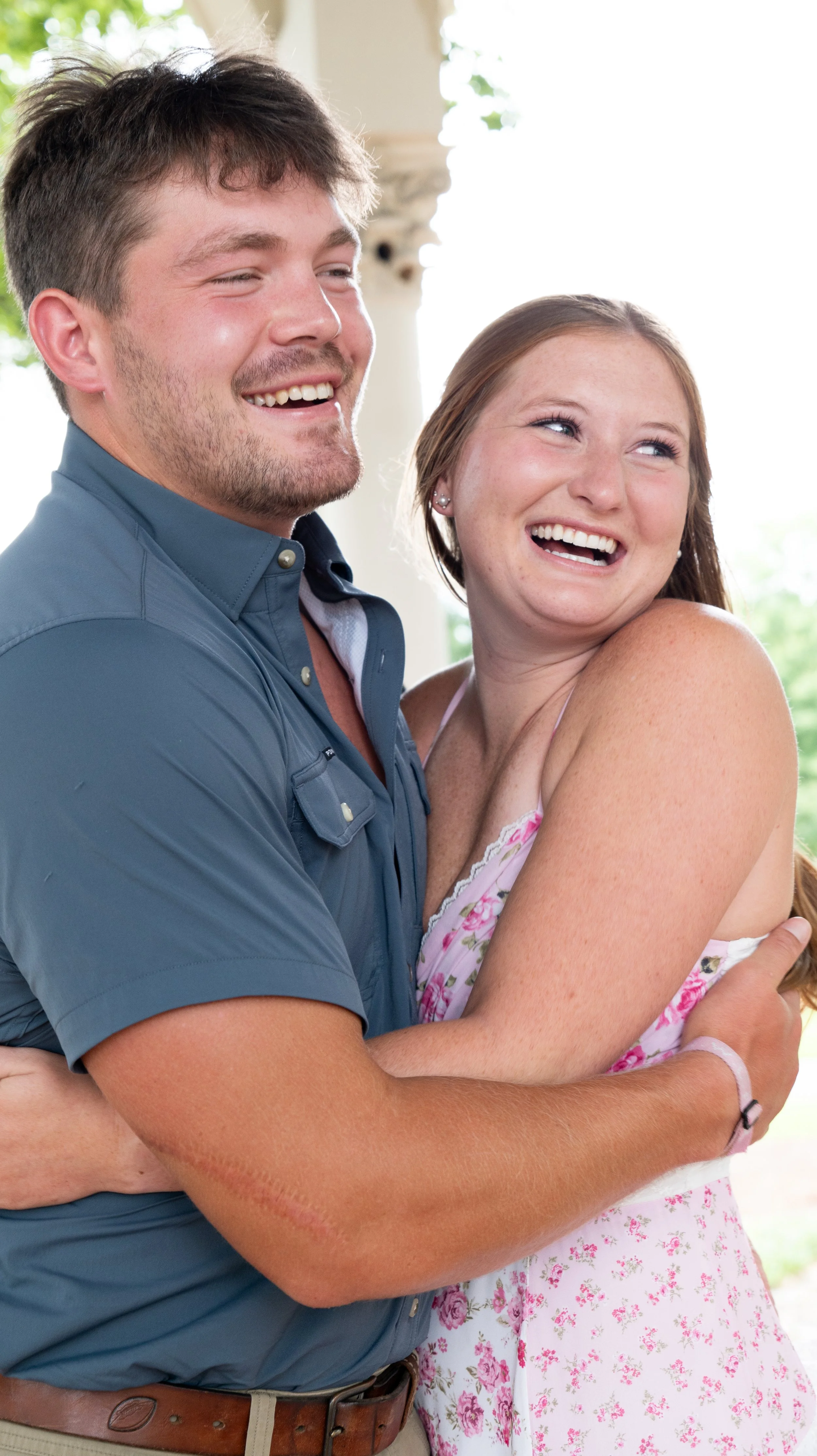 A man and woman happily hugging each other indoors with natural light and greenery outside. The man has short brown hair, a beard, and is wearing a blue shirt, while the woman has long red hair and is wearing a pink floral dress.