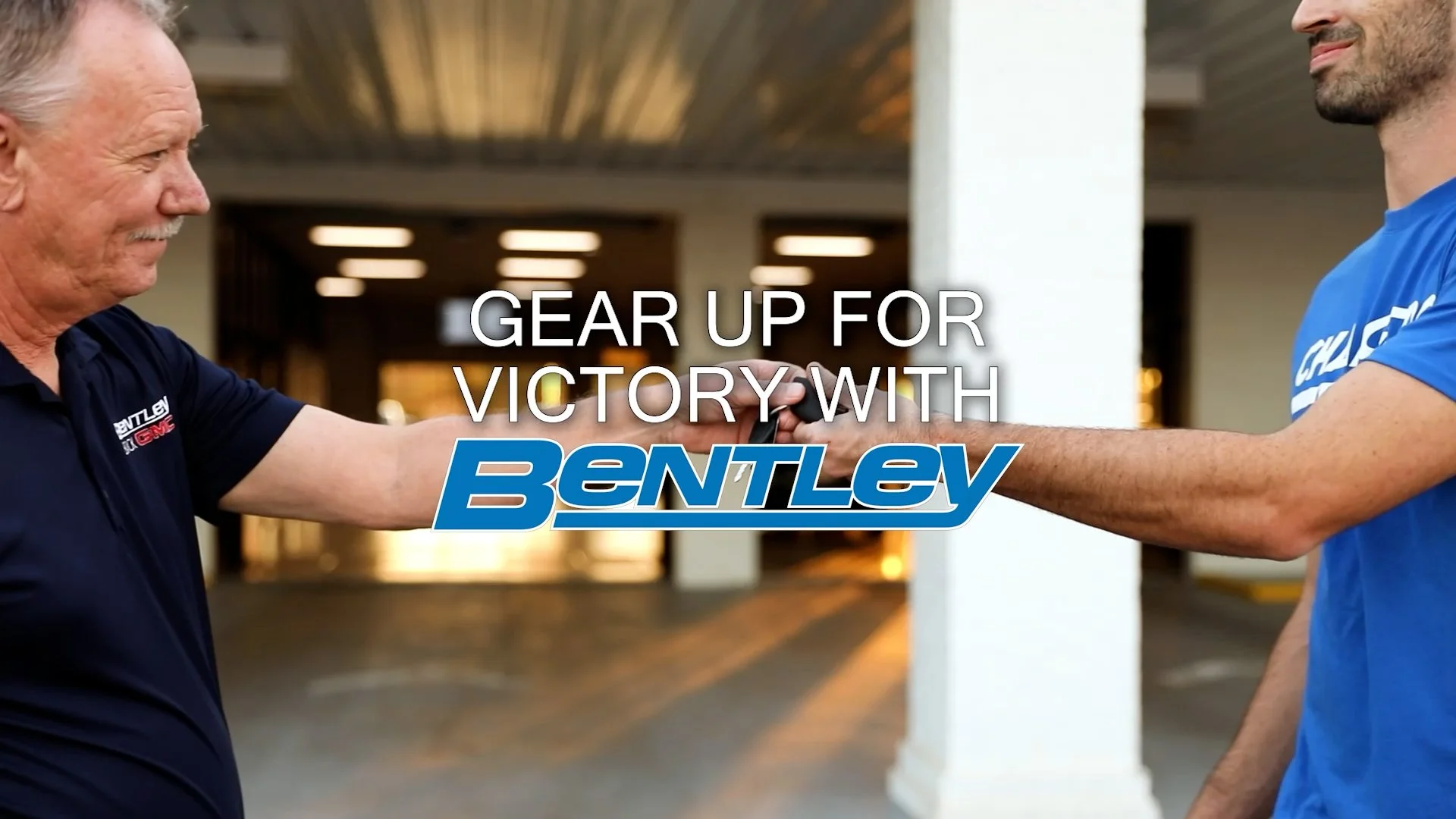 An older man and a younger man shake hands, both wearing Bentley branded clothing, standing outdoors in front of a building with a high ceiling.