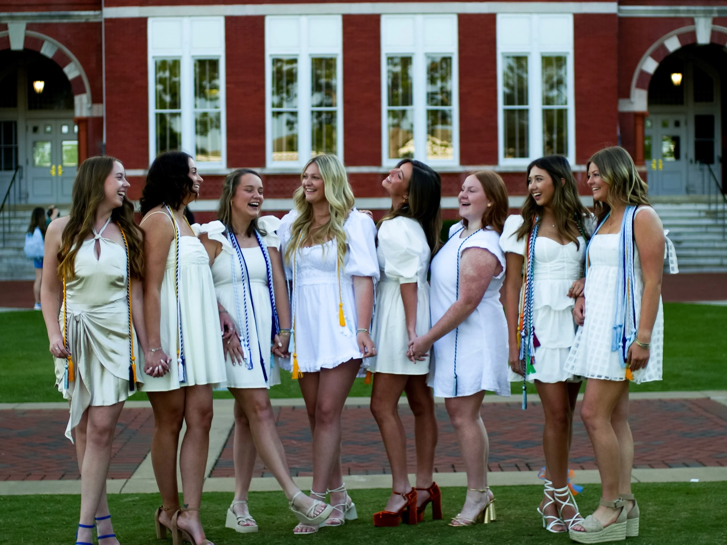 A group of eight women dressed in white dresses, holding hands, and wearing graduation stoles, standing outside in front of a red brick building with large windows during daytime.