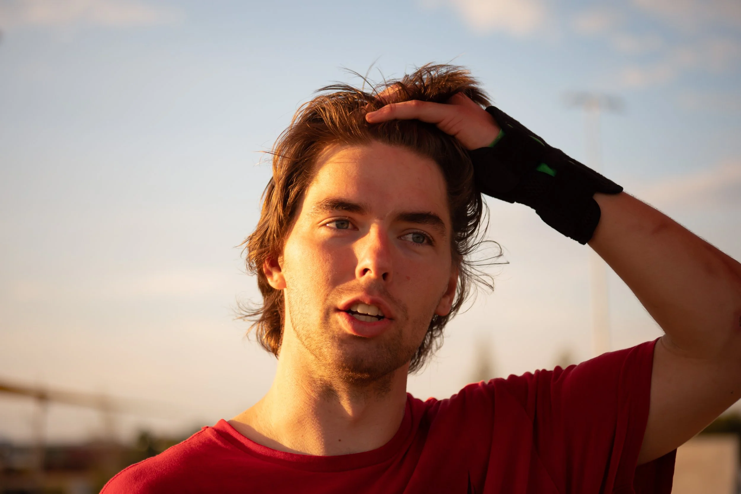 A young man with light brown hair, wearing a red shirt, touching his head with one hand, outdoors during sunset under a blue sky with clouds.