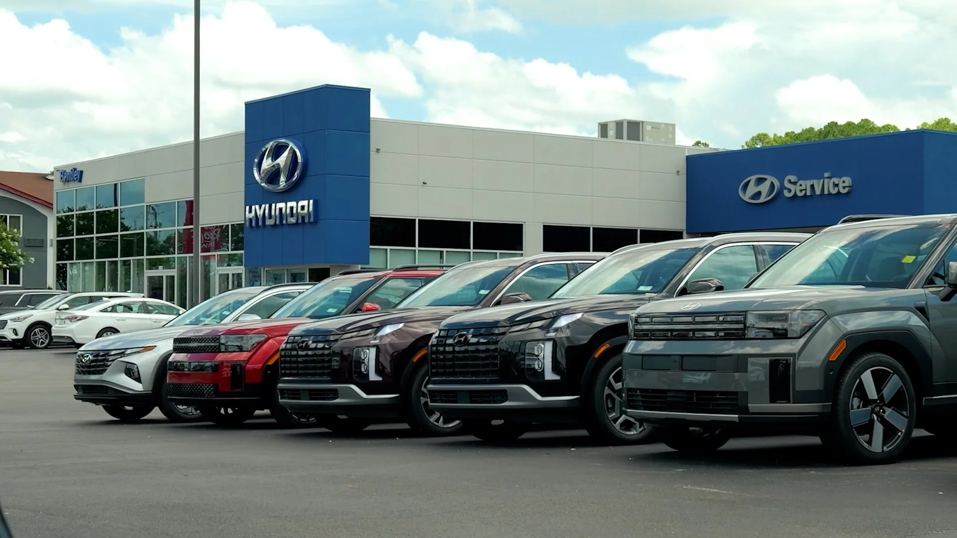 Car dealership with multiple new Hyundai vehicles parked in front and a Hyundai service center building in the background.