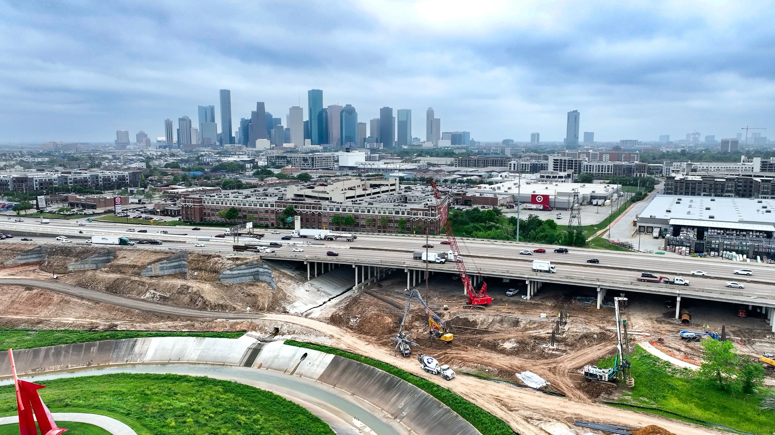 A city skyline with tall skyscrapers under a cloudy sky, viewed from a highway under construction with machinery and soil erosion in the foreground.