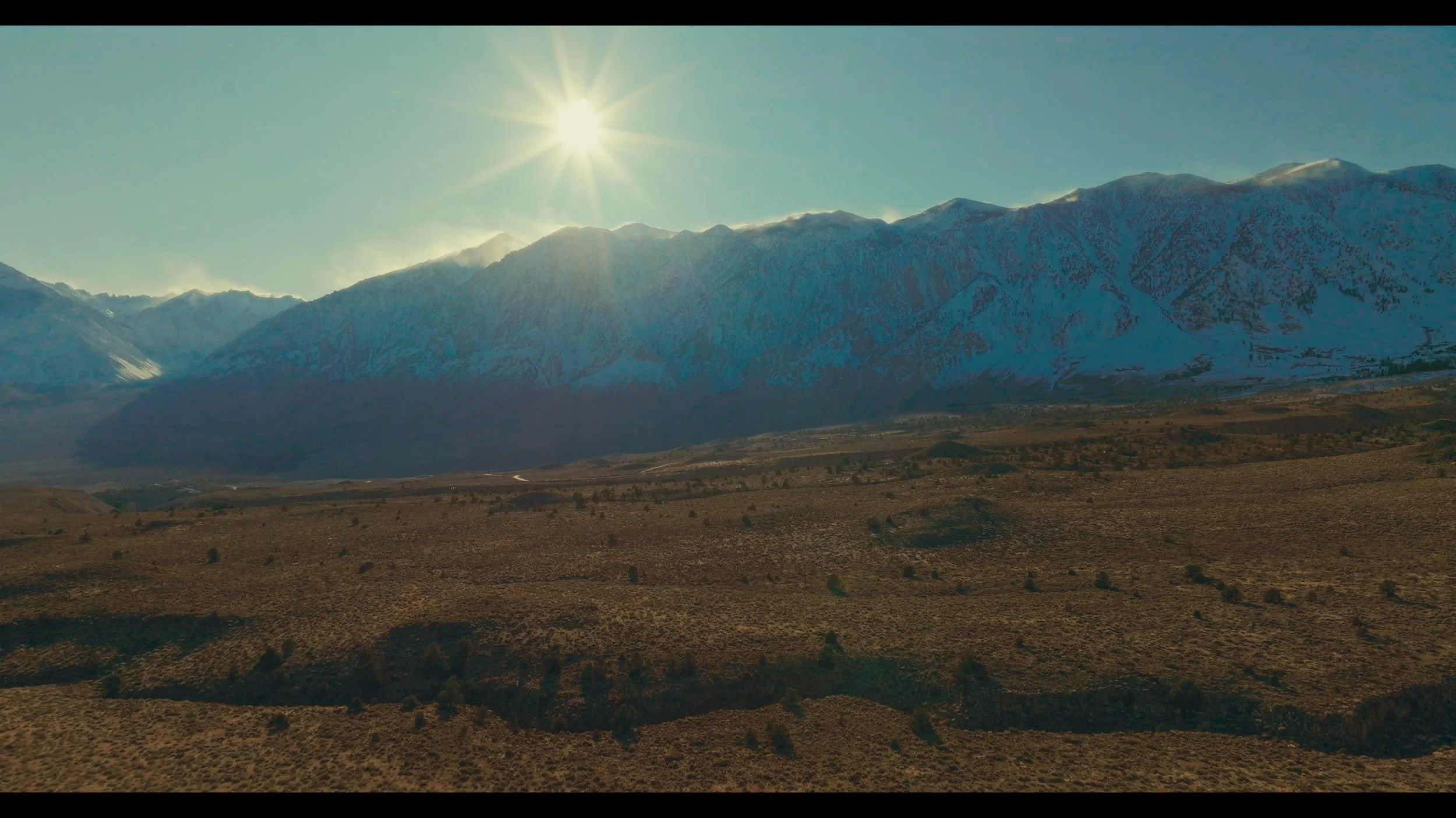 Scenic view of a mountain range with snow-capped peaks under a bright sun, dry brown terrain in the foreground