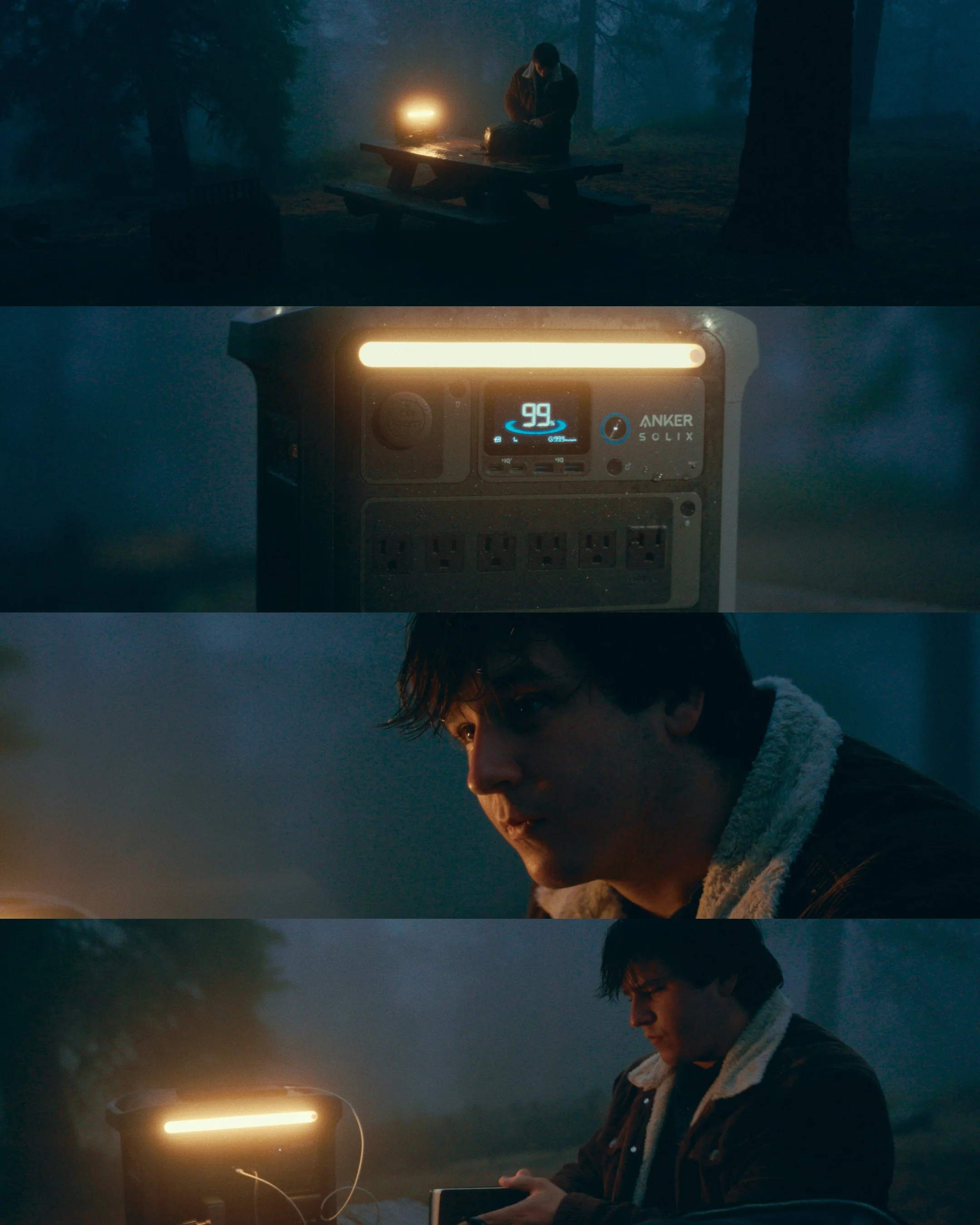 A young man sitting at a picnic table outdoors at night, with a lantern and a portable power station nearby, looking at his phone.