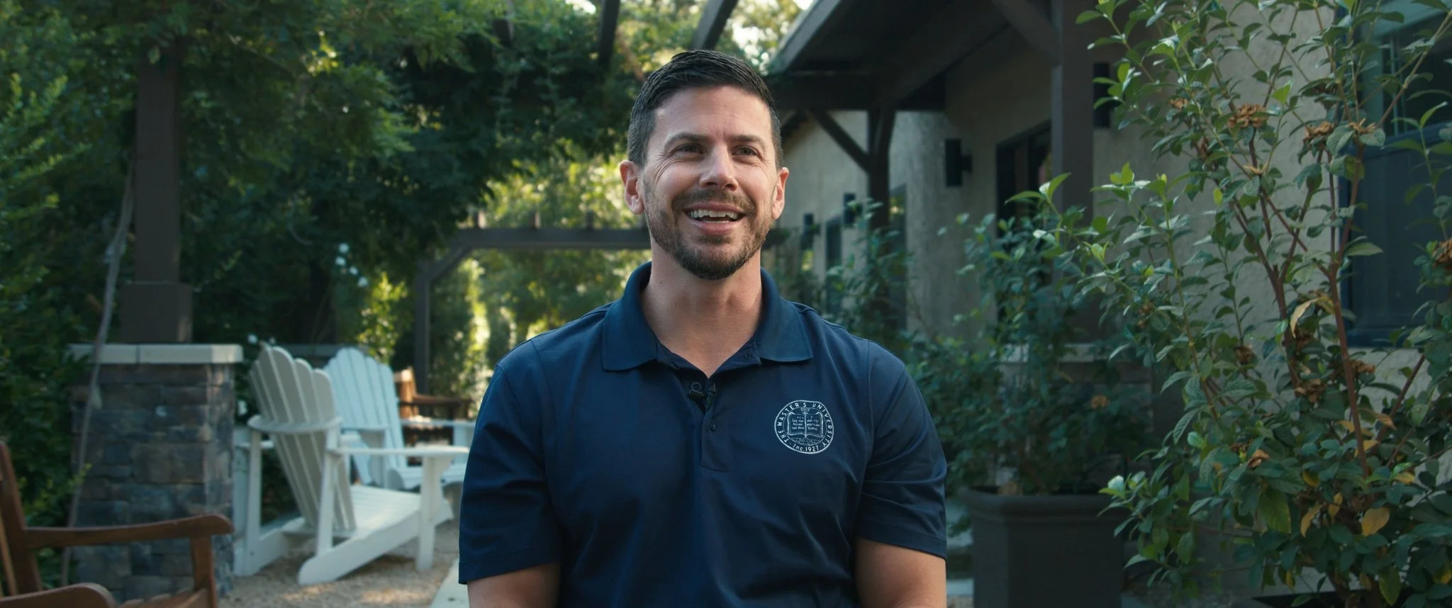 Man smiling outdoors in a garden with trees, bushes, and outdoor furniture.