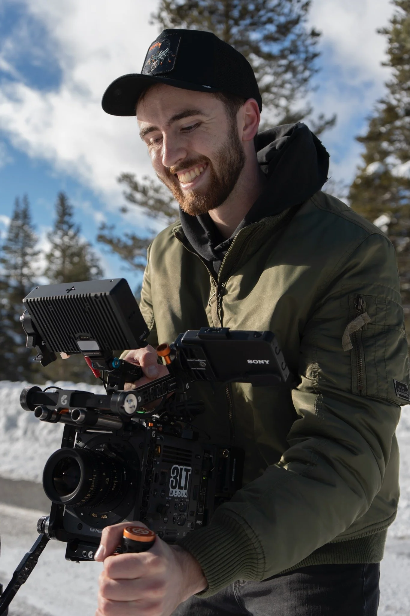 A man wearing a black cap, green jacket, and black hoodie smiling while operating professional filmmaking equipment outdoors on a snowy day with trees and a blue sky in the background.