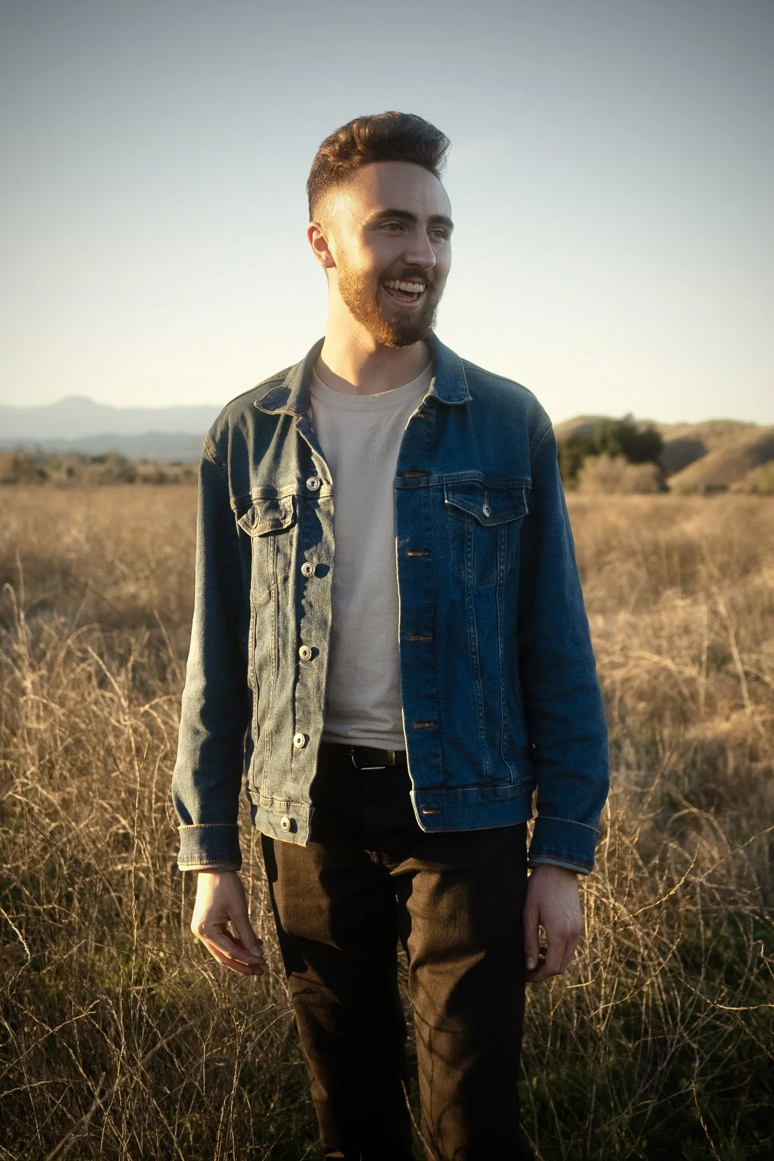 A man with dark, curly hair, glasses, a nose ring, and a beard wearing a dark green sweater sits indoors.