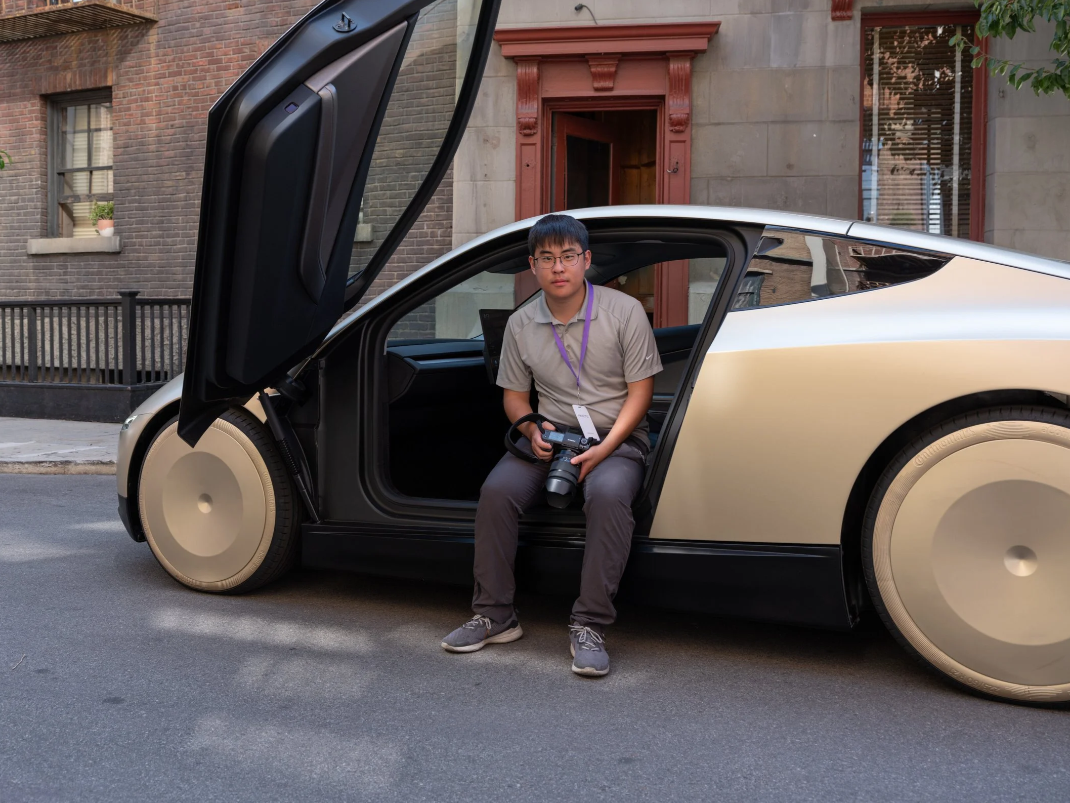 Young man sitting in an autonomous vehicle with gull-wing door open, holding a camera, on urban street.