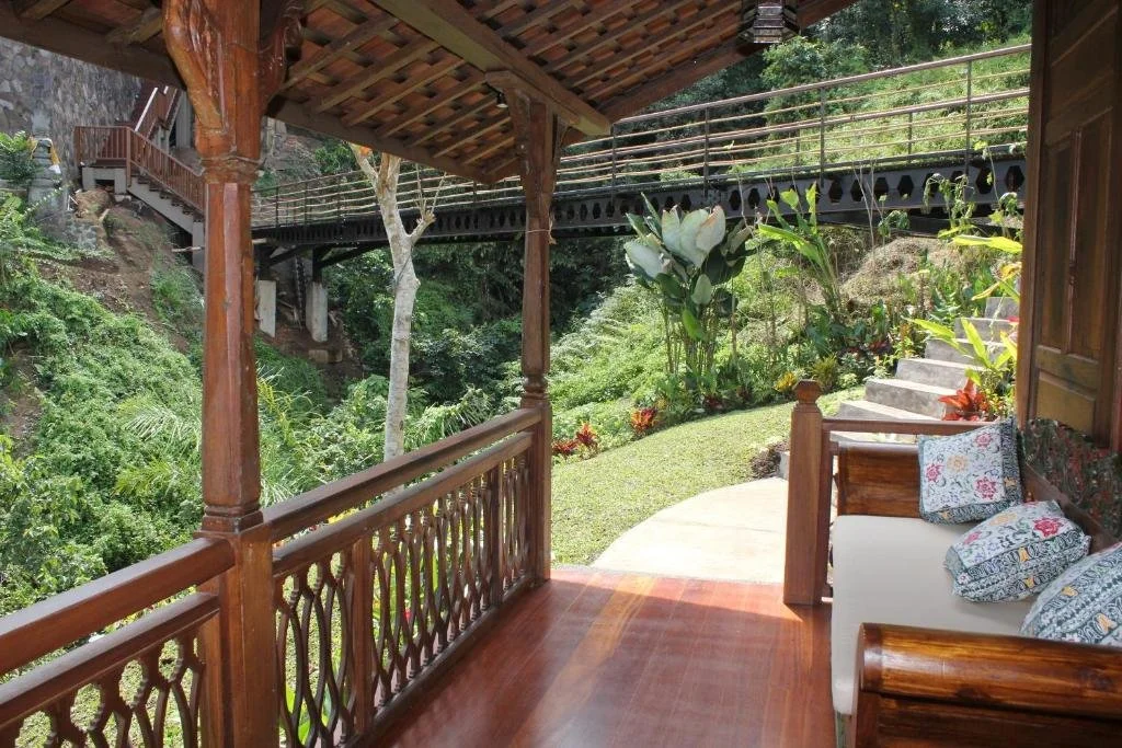 View from a wooden porch with a white sofa and patterned cushions, overlooking a lush garden with stairs and a bridge in the background.