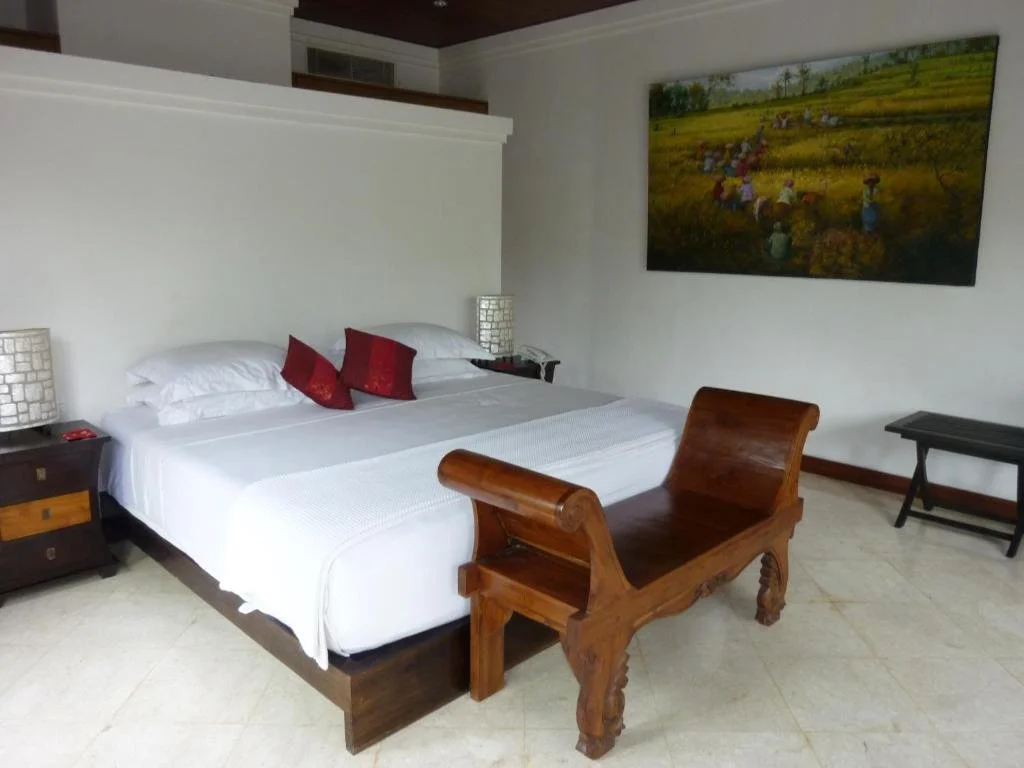 A hotel room with a bed, two nightstands, a painting of a rice field, and a wooden bench.