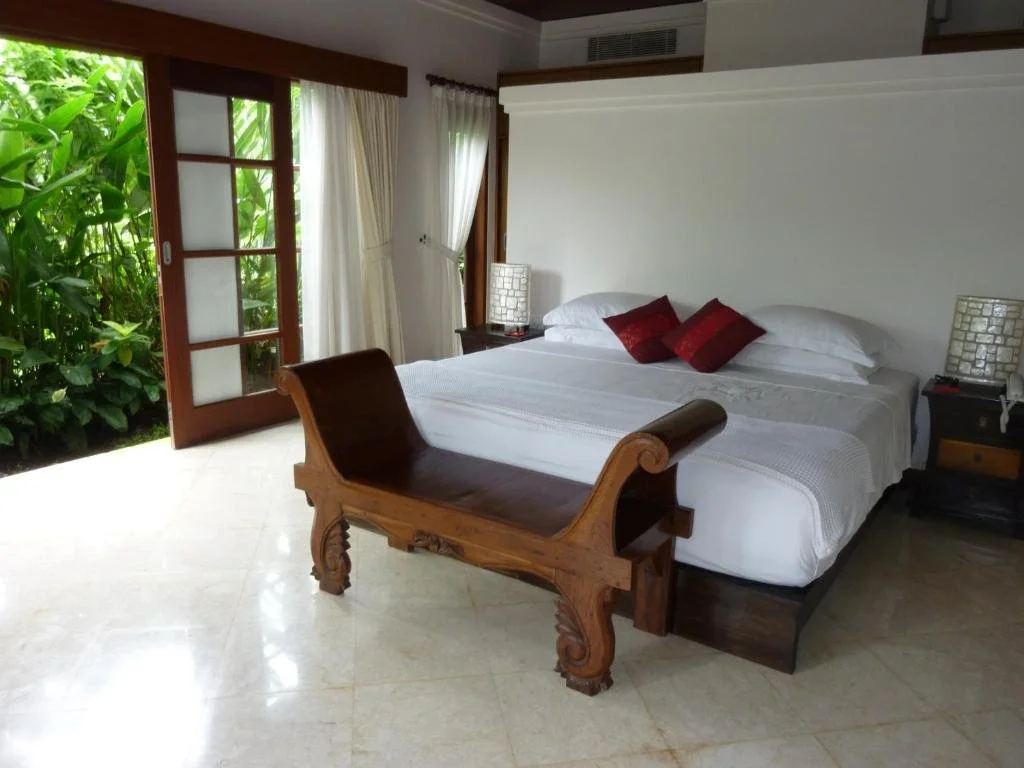 A hotel room with a large bed, white bedding, and red pillows. There are two nightstands with lamps, and a wooden bench at the foot of the bed. The room has large glass doors leading to an outside garden with green plants.
