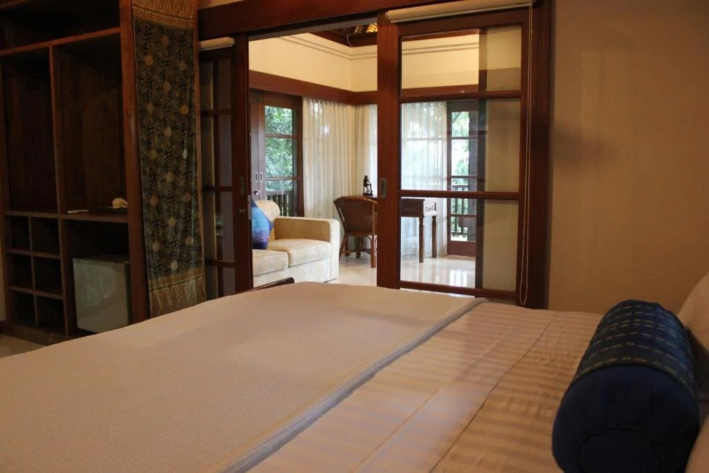Hotel room with a bed, wooden shelves, and a view into a living area with a sofa, chair, and glass sliding doors leading to a balcony, with curtains and greenery outside.