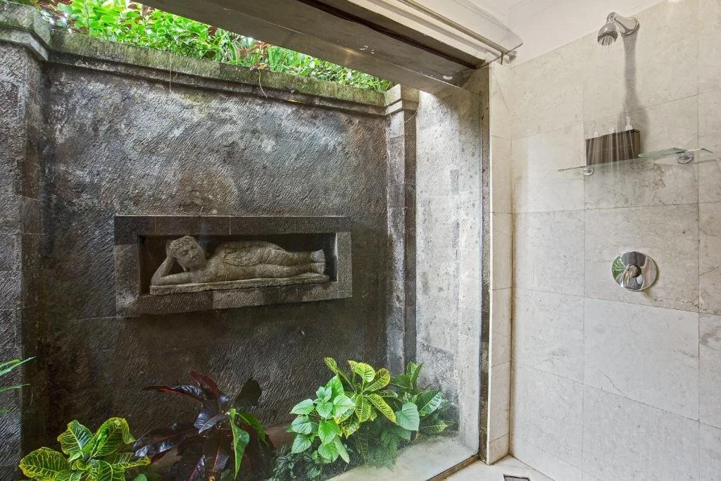 Indoor shower with a large glass window showing an outdoor stone wall with a reclining Buddha statue and green plants.