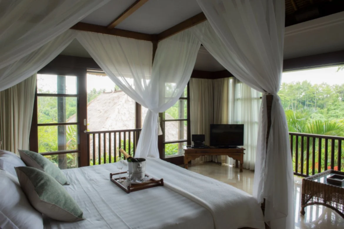 A bedroom with a four-poster bed, sheer white curtains, green pillows, a small table with a tray, a TV, and large windows showing a lush tropical view outside.