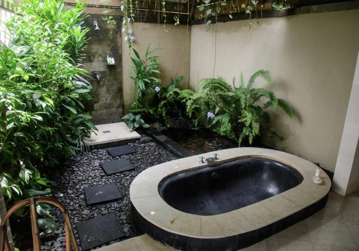A small indoor garden with a bathtub, lush green plants, a pebble pathway, and a water feature.