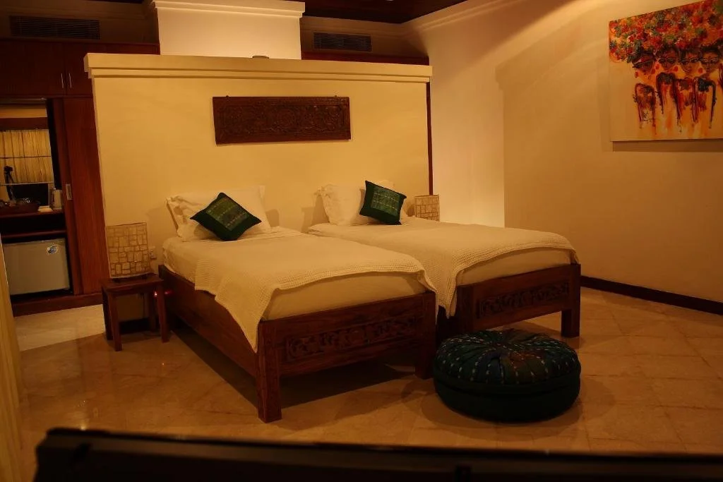 Two neatly made twin beds with white bedding and green accent pillows, in a warmly lit hotel room with wooden furniture, a painting on the wall, a small round ottoman, and a side table.