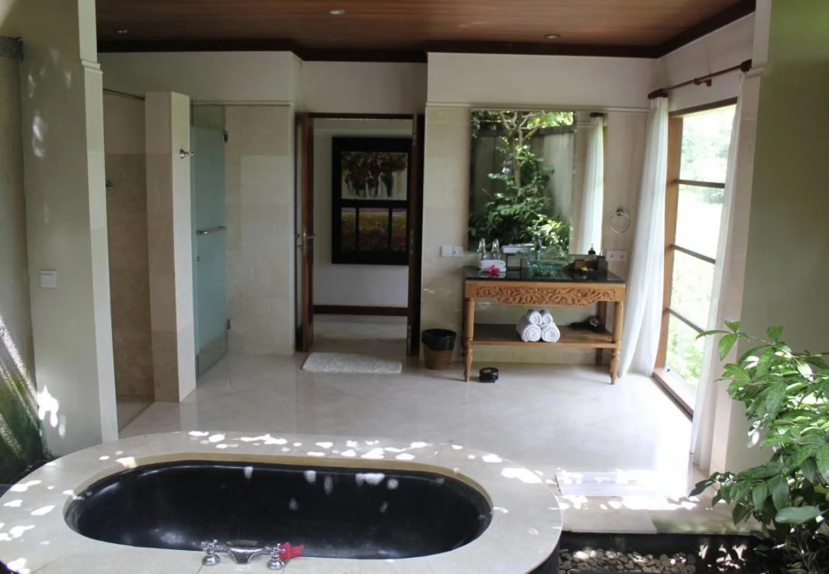 Bathroom with a large black soaking tub, wooden ceiling, and natural light from windows with white curtains. A wooden table holds rolled white towels and toiletries, overlooking a garden view.