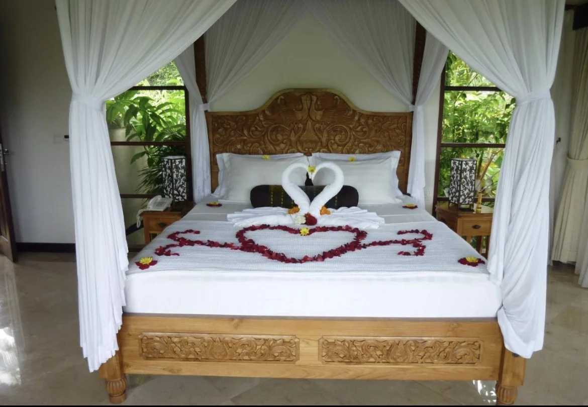 Decorated hotel bed with white sheets, bed towels folded into swans facing each other, forming a heart shape, surrounded by flower petals on the bed and on pillows, with lush green foliage outside the windows.