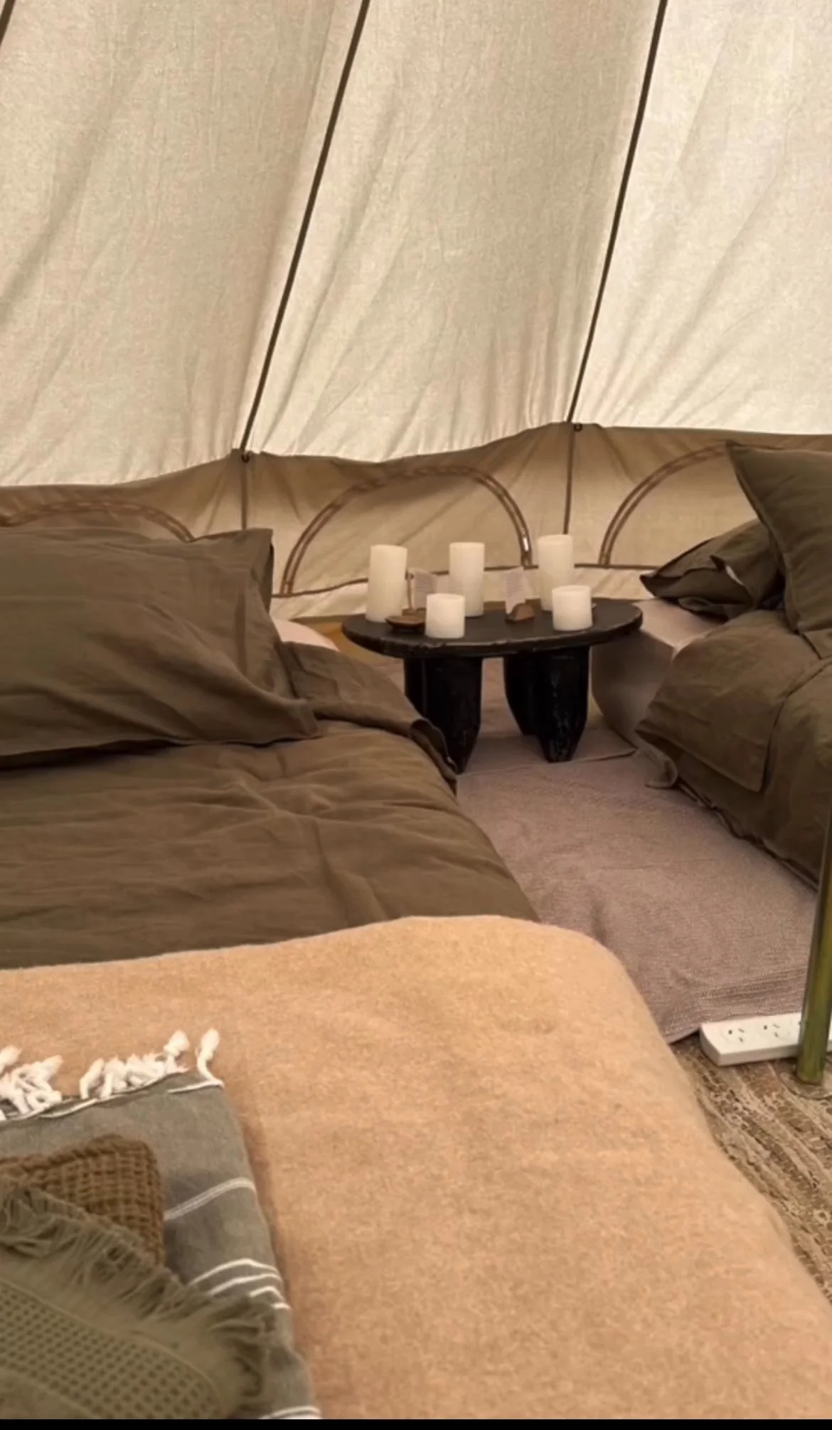 Inside a cozy tent with beige walls, two beds with brown and beige bedding, and a black round table in between holding multiple white candles.